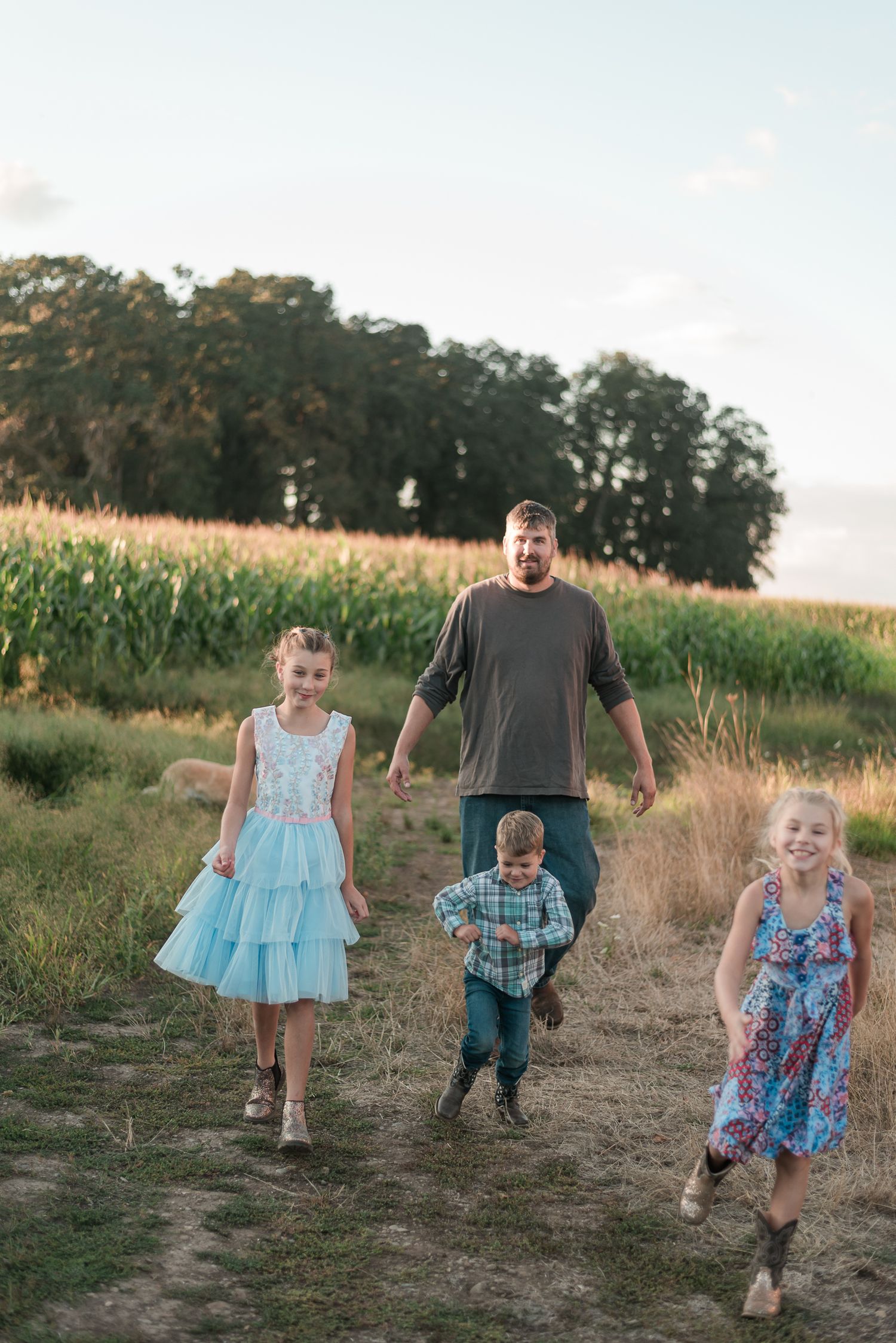 Dad and three kids laughing and walking together along a rural dirt path beside a green cornfield.