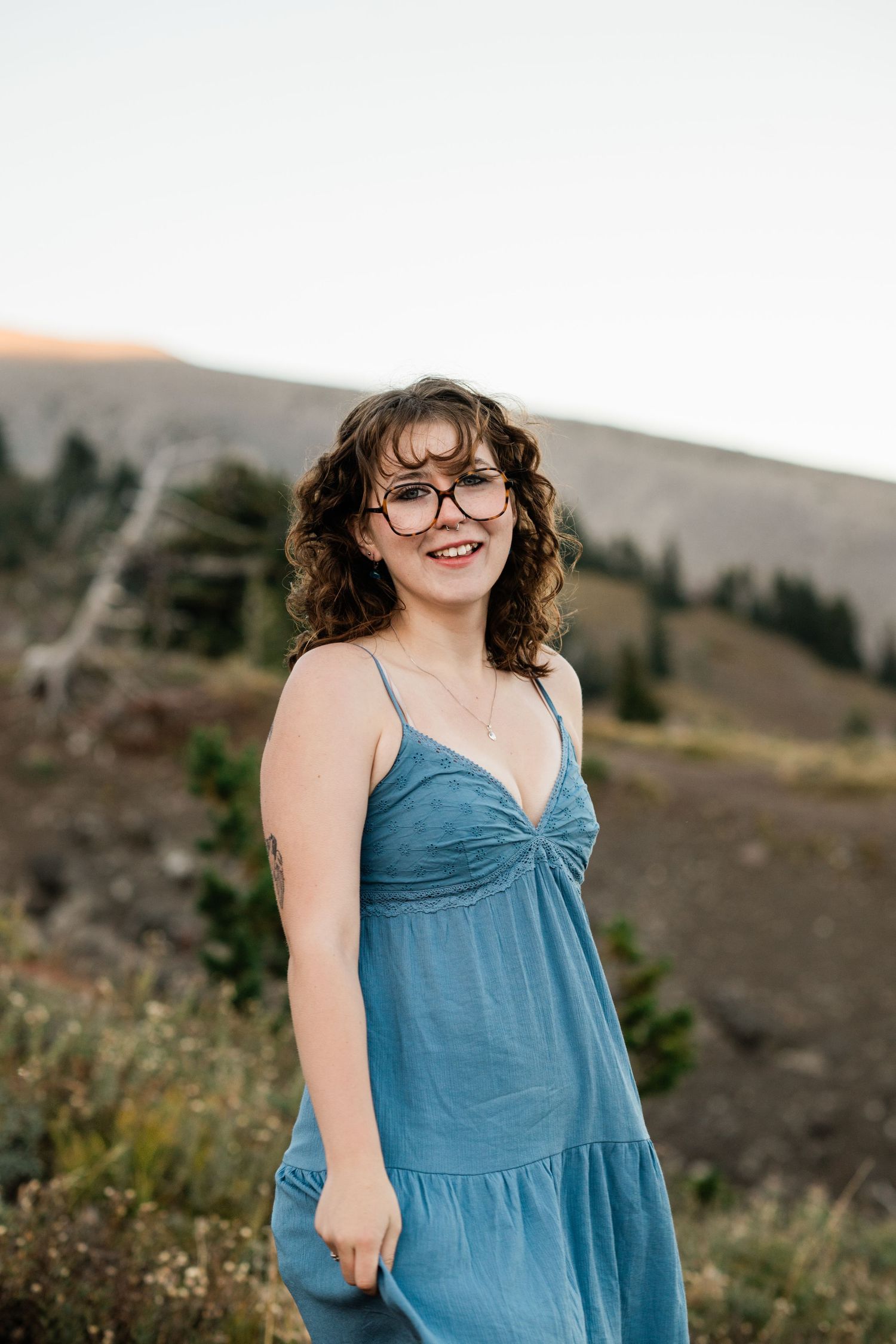 Person in a teal summer dress poses outdoors against a scenic mountain landscape at sunset.
