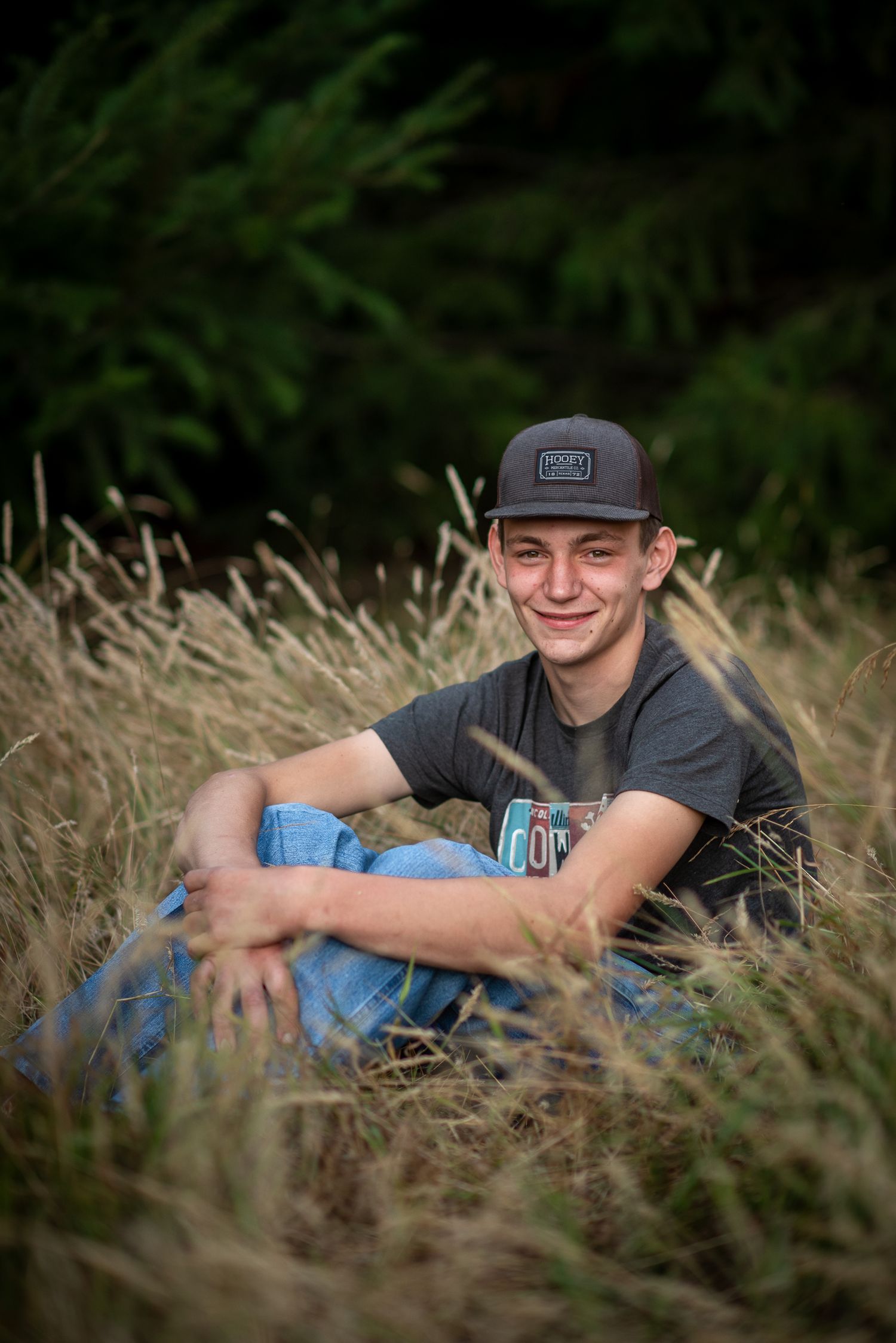 Smiling teenage boy in cap and jeans sitting relaxed in tall golden grass outdoors.