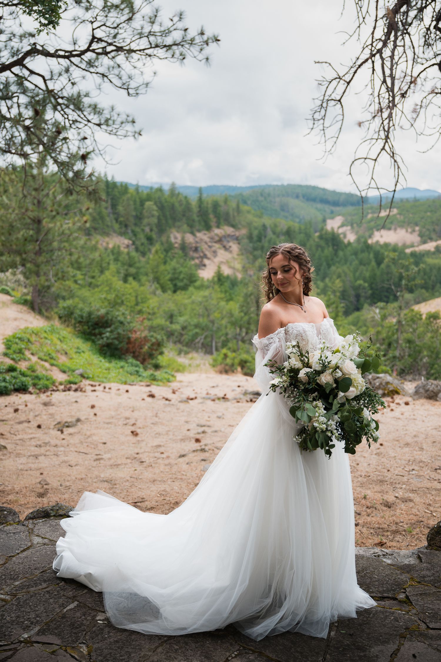 Elegant bride in off-shoulder tulle gown holds white bouquet with scenic mountain forest backdrop.