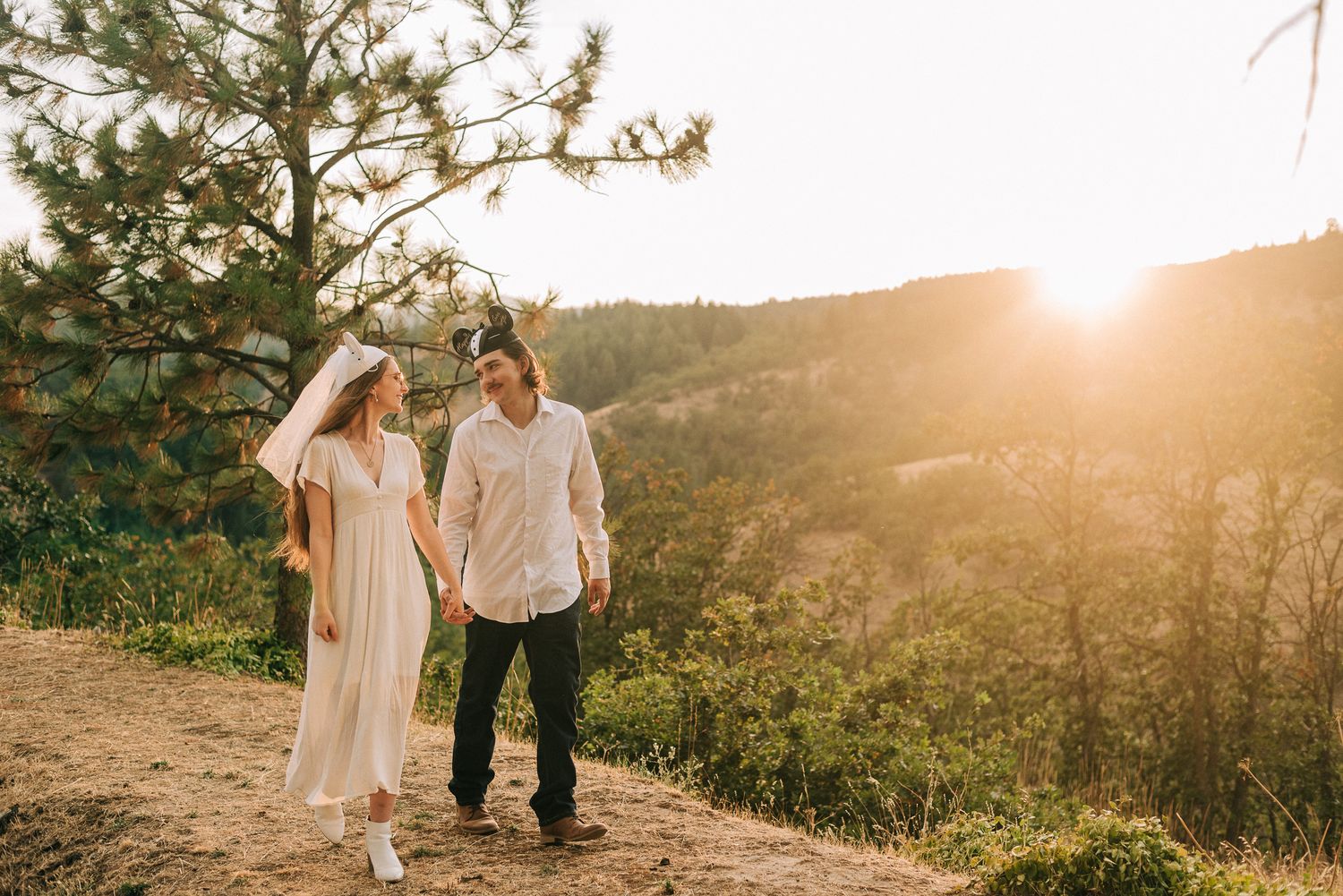 A newlywed couple walks hand in hand along a scenic overlook at golden sunset.