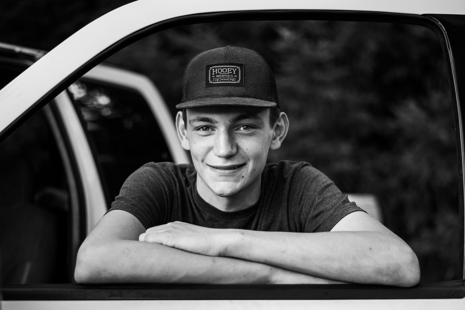 Smiling teenage boy in a cap leaning on a white truck window in black and white photo.