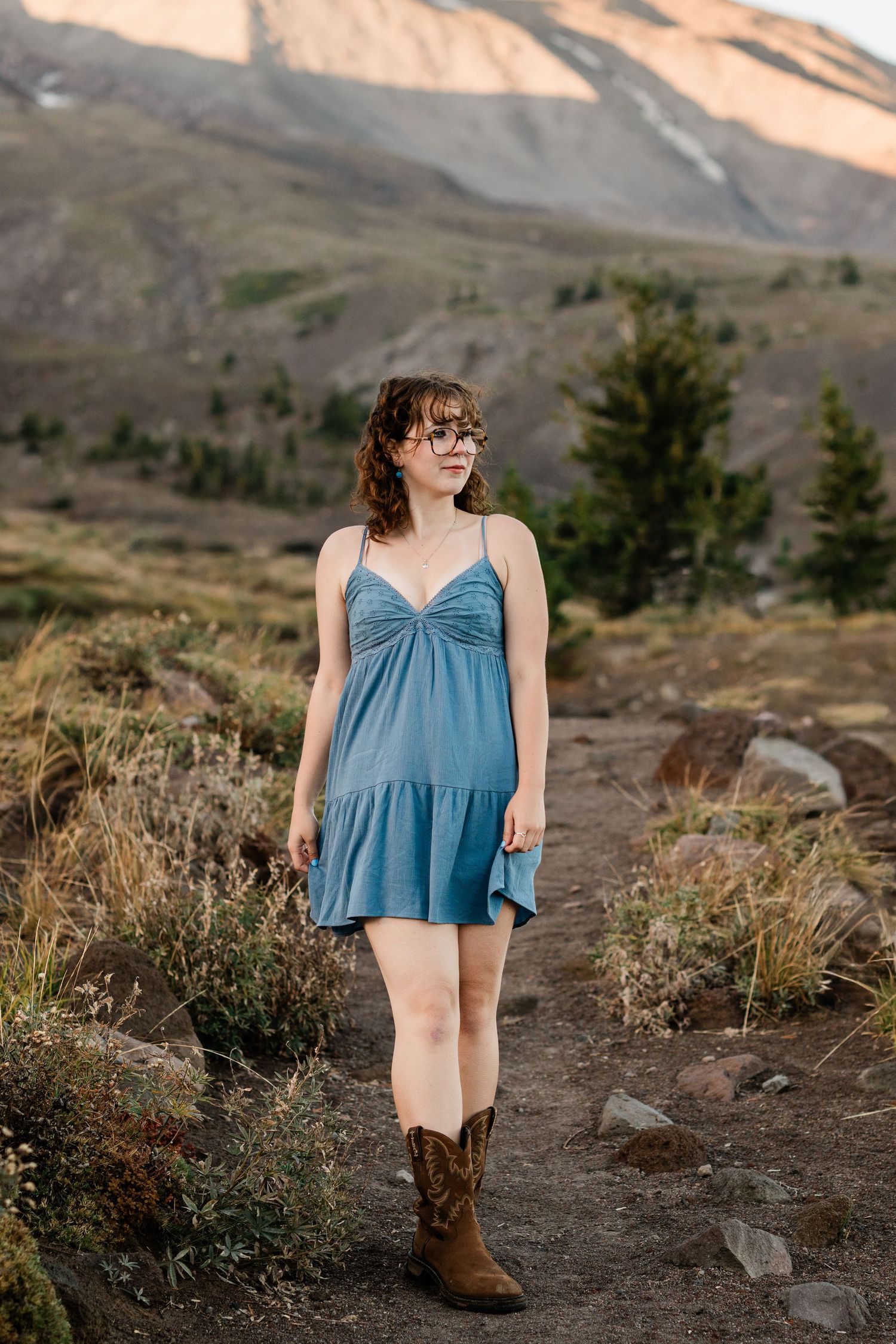 A person in a blue top stands on a mountain trail surrounded by rugged terrain and wildflowers at sunset.