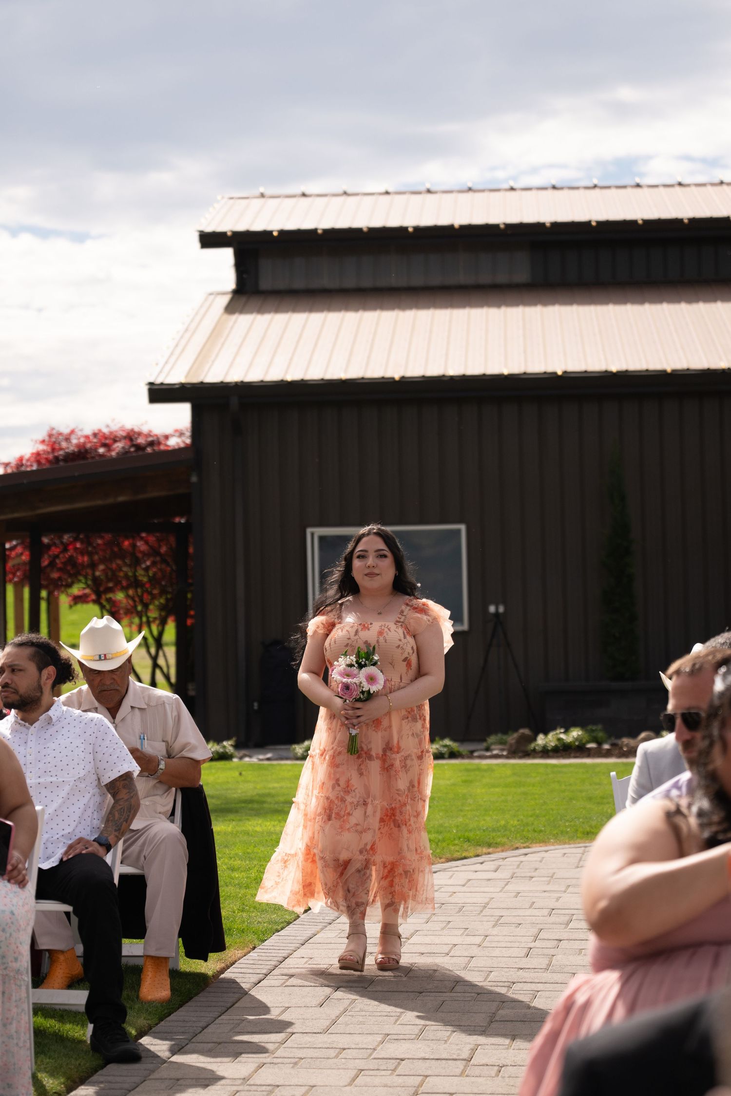 Bridesmaid in floral peach dress walks down outdoor wedding aisle holding bouquet, guests seated beside dark barn venue.