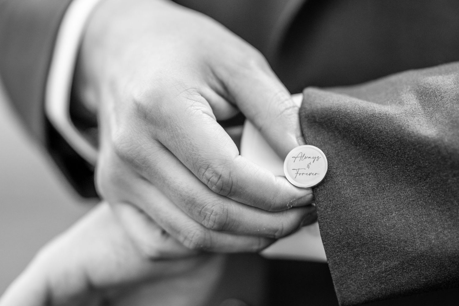Close-up black and white photo of groom fastening engraved cufflinks on wedding suit jacket sleeve.