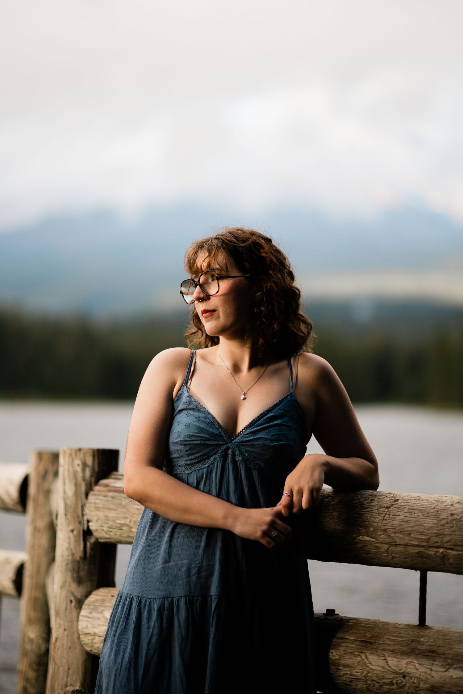 A person in a blue dress leans against a wooden railing by a serene lake with mountains in the background.
