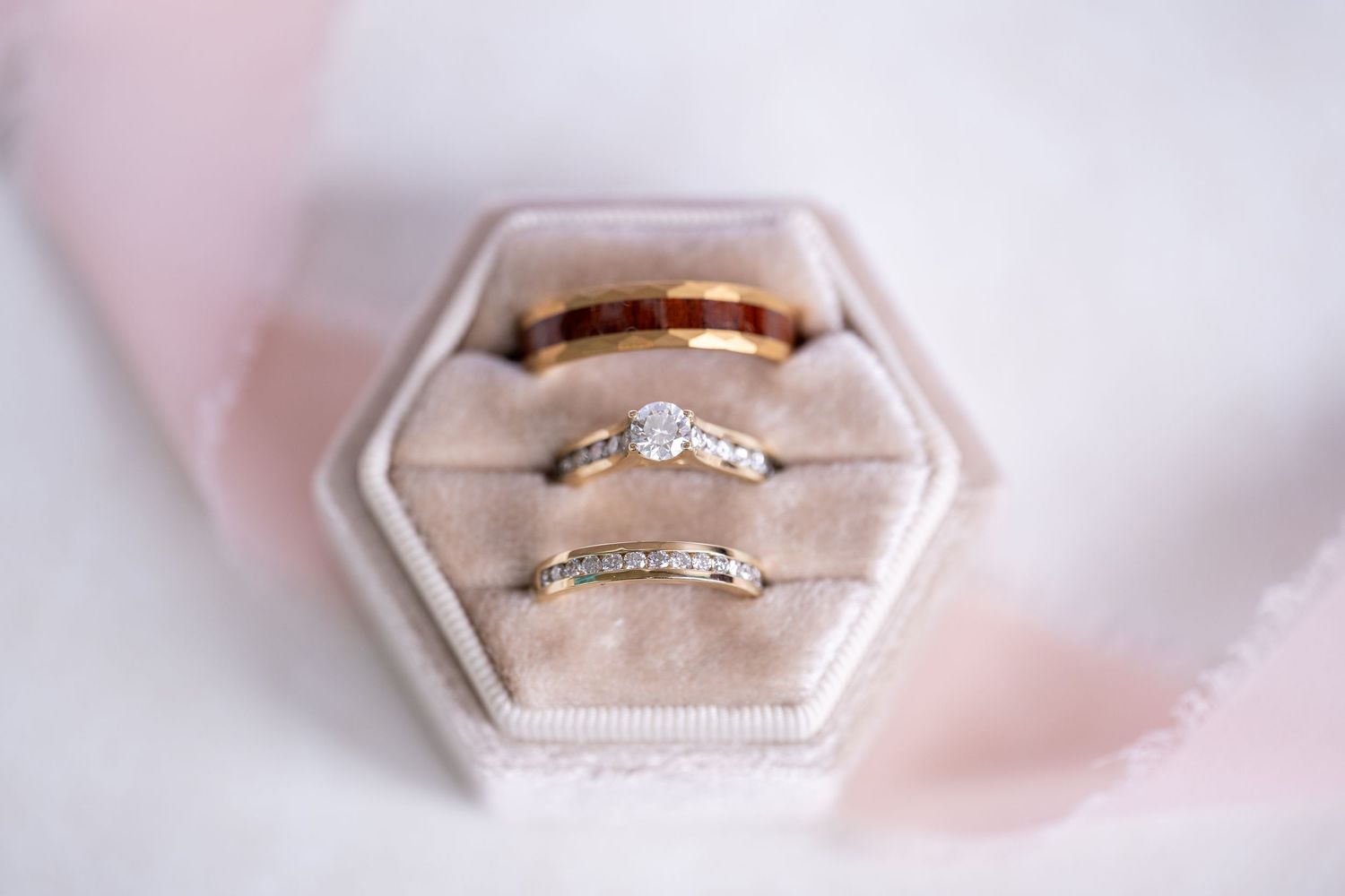 Three wedding rings displayed elegantly in a hexagonal velvet ring box with soft pink ribbon backdrop.
