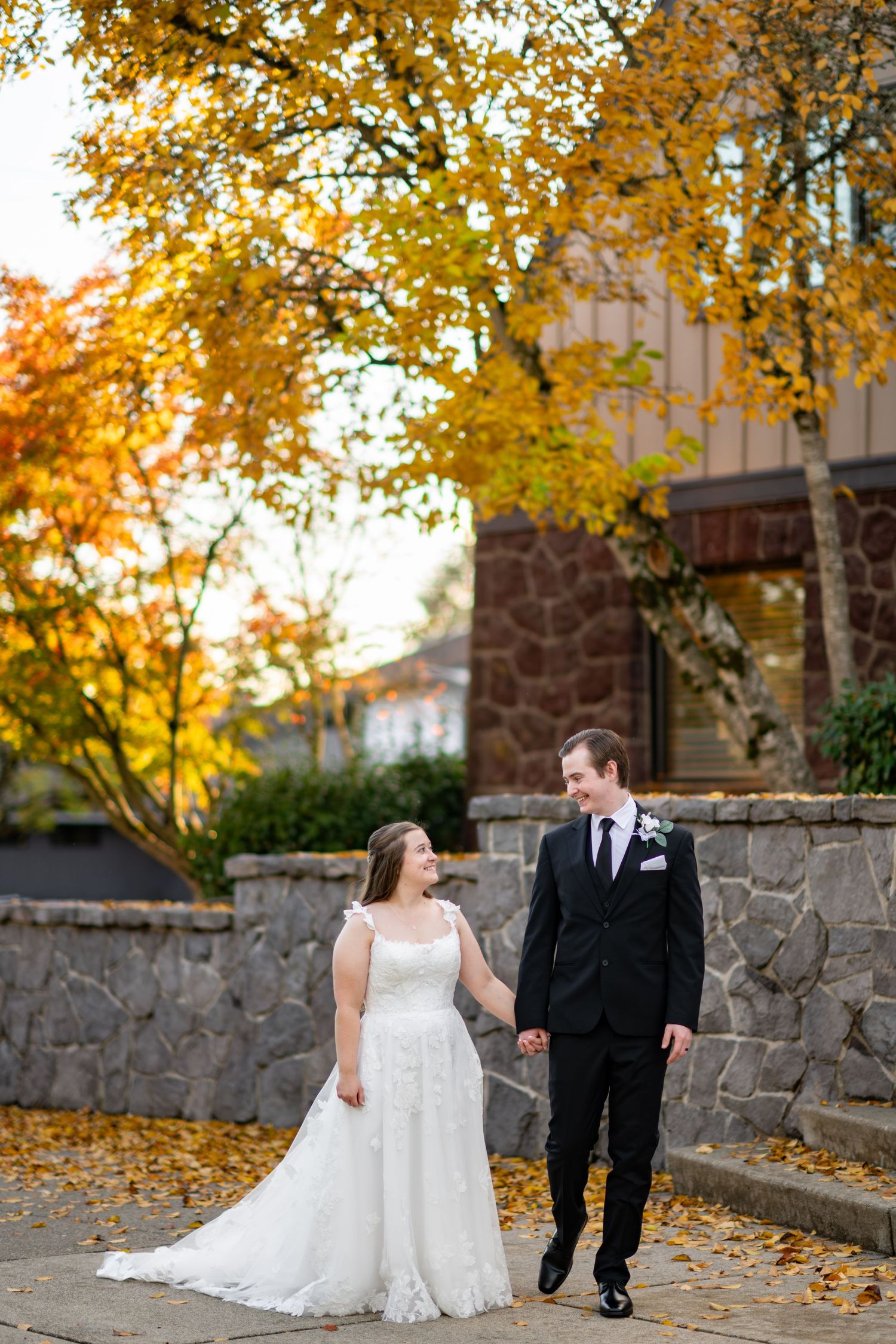 Bride and groom walk hand-in-hand among vibrant autumn foliage beside a stone building at their fall wedding.