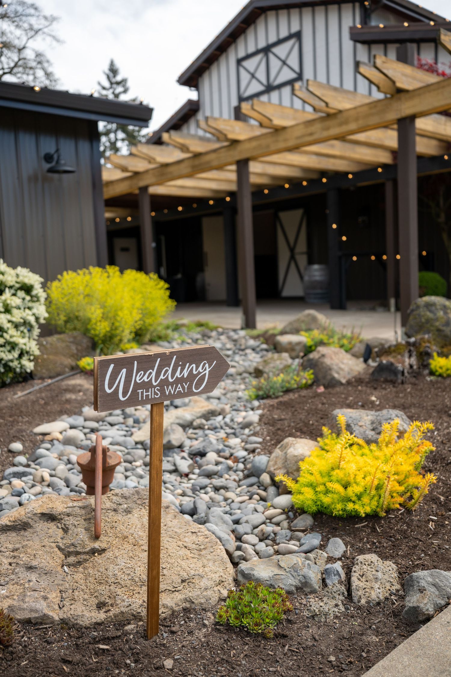 Rustic barn wedding venue with a 'Wedding This Way' sign, pergola with string lights, and landscaped rock garden.