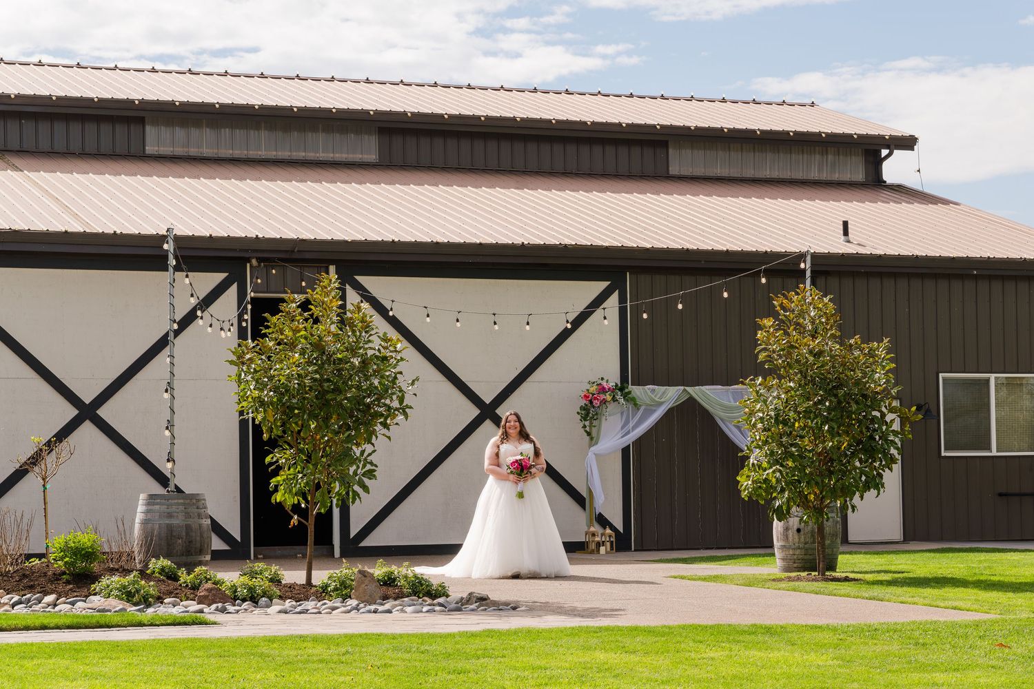 Bride in white gown poses outside a rustic barn wedding venue with string lights and lush green landscaping.
