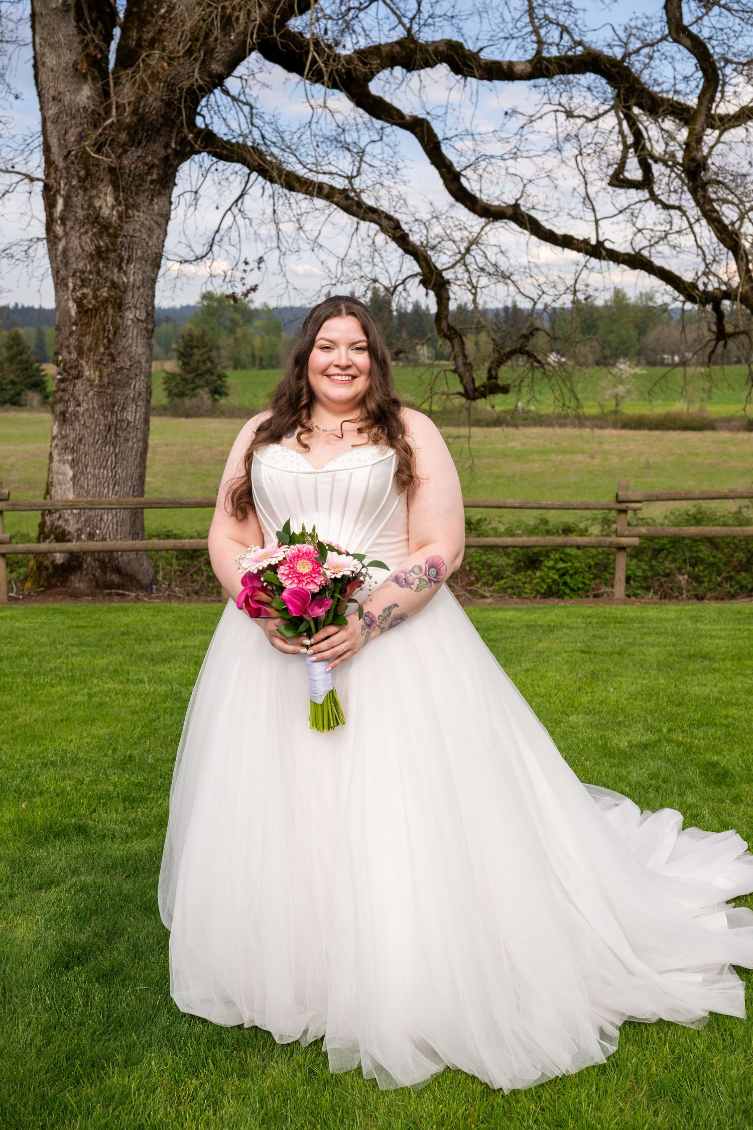 Smiling bride in white tulle gown holding pink bouquet, standing on green lawn with wooden fence and trees.