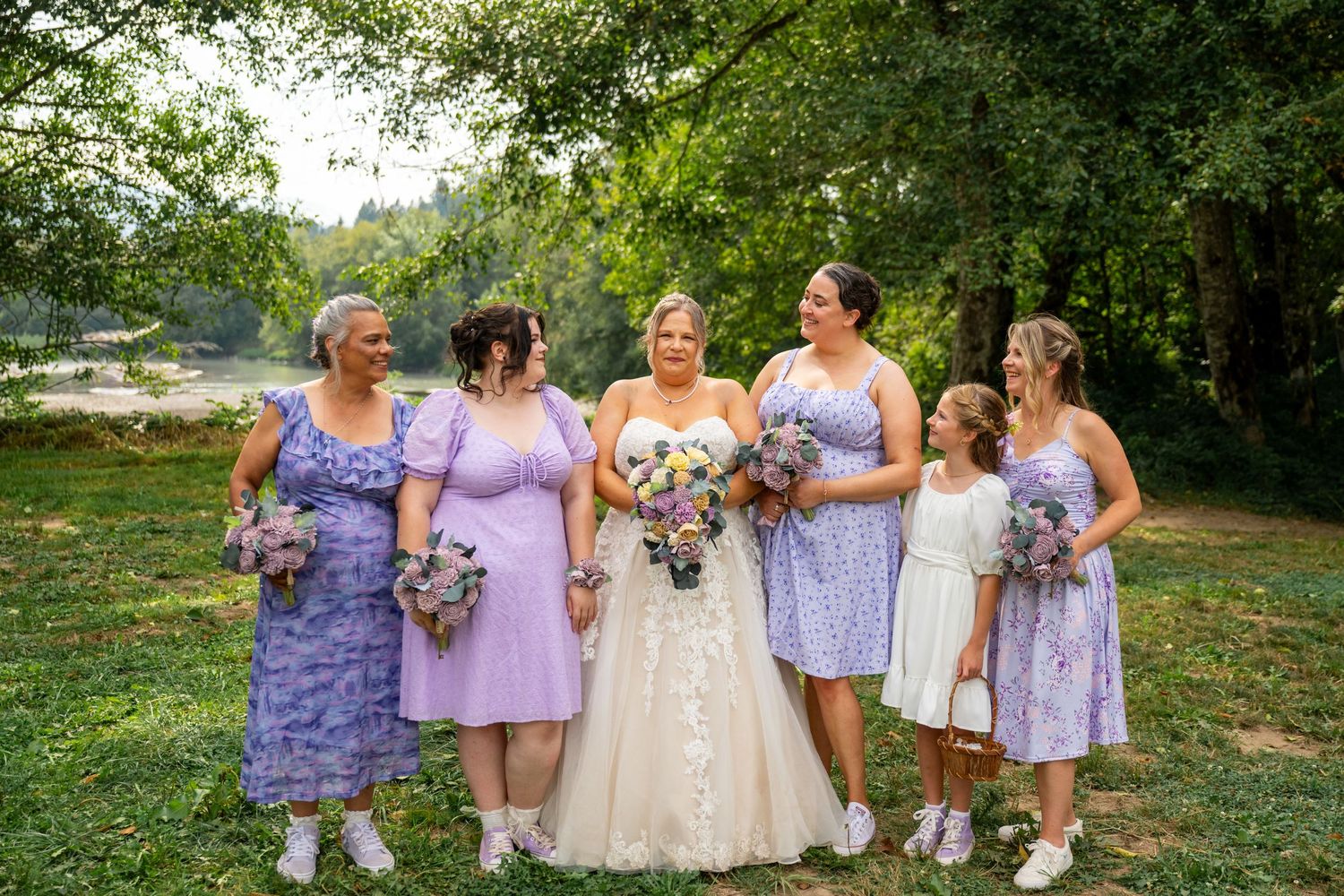 Wedding party in purple dresses posing outdoors under green trees.