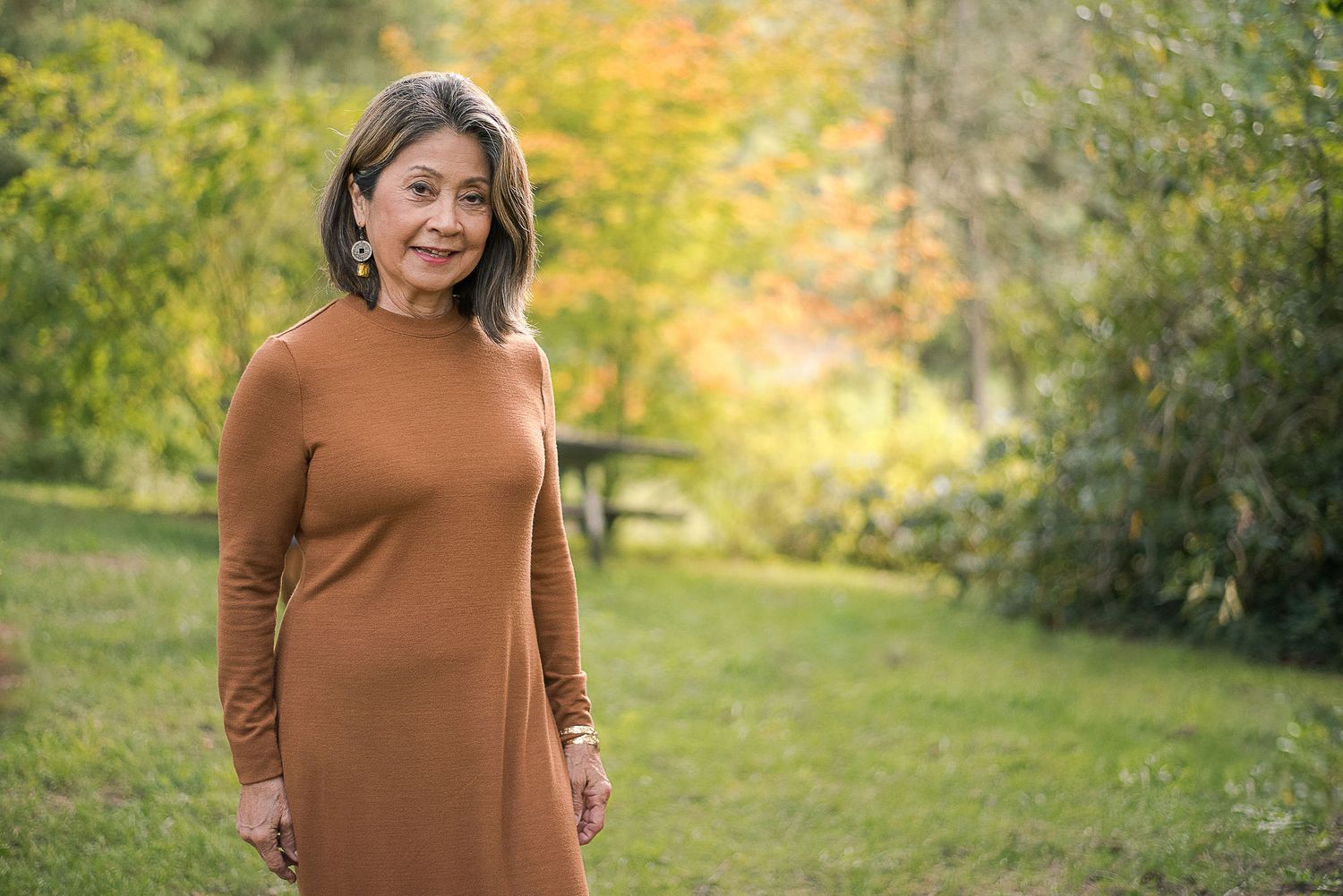 Woman in a brown turtleneck sweater poses outdoors against an autumn backdrop.