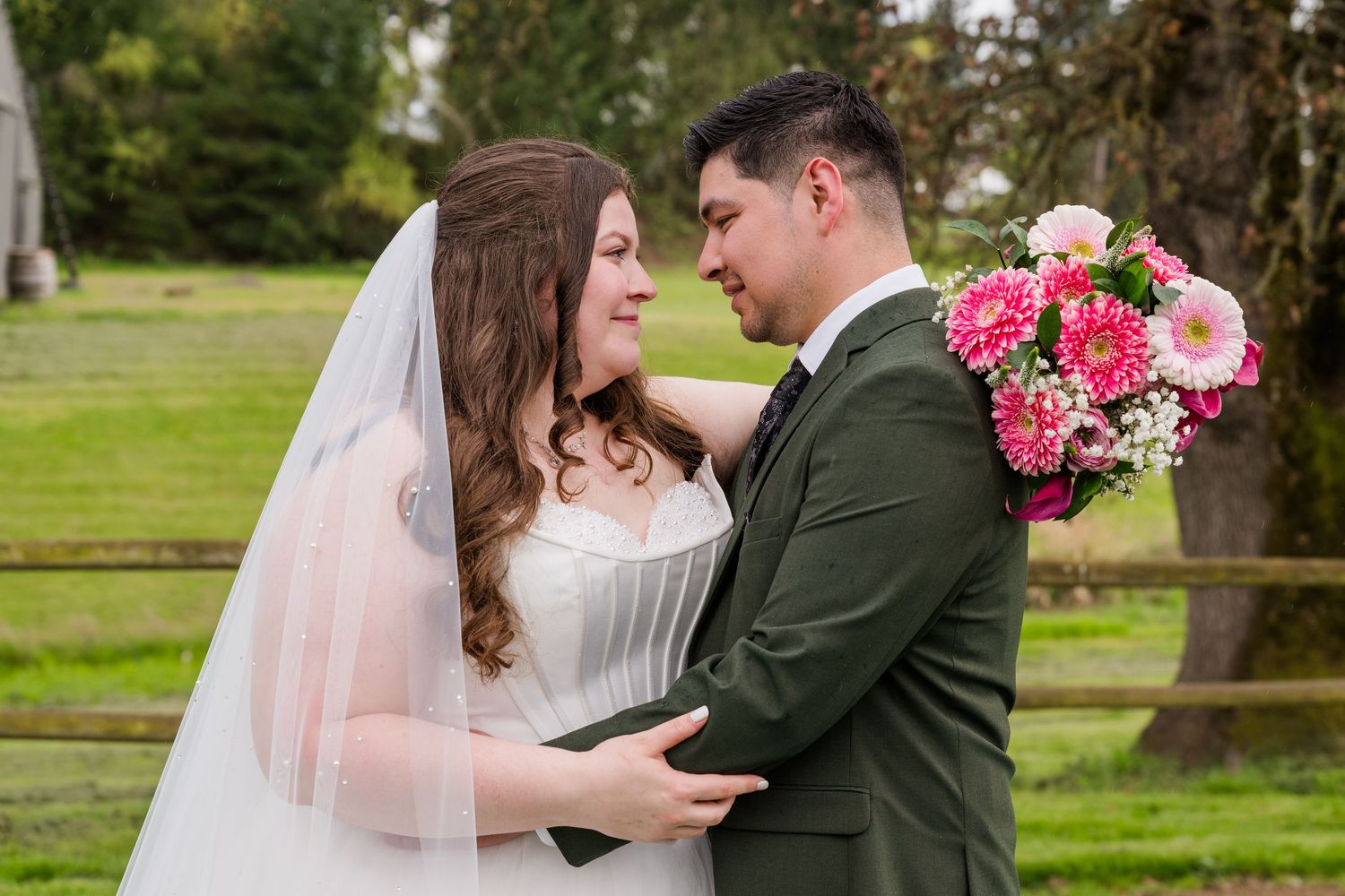 Bride and groom share an intimate moment, with the groom in an olive suit holding pink flowers at an outdoor wedding.