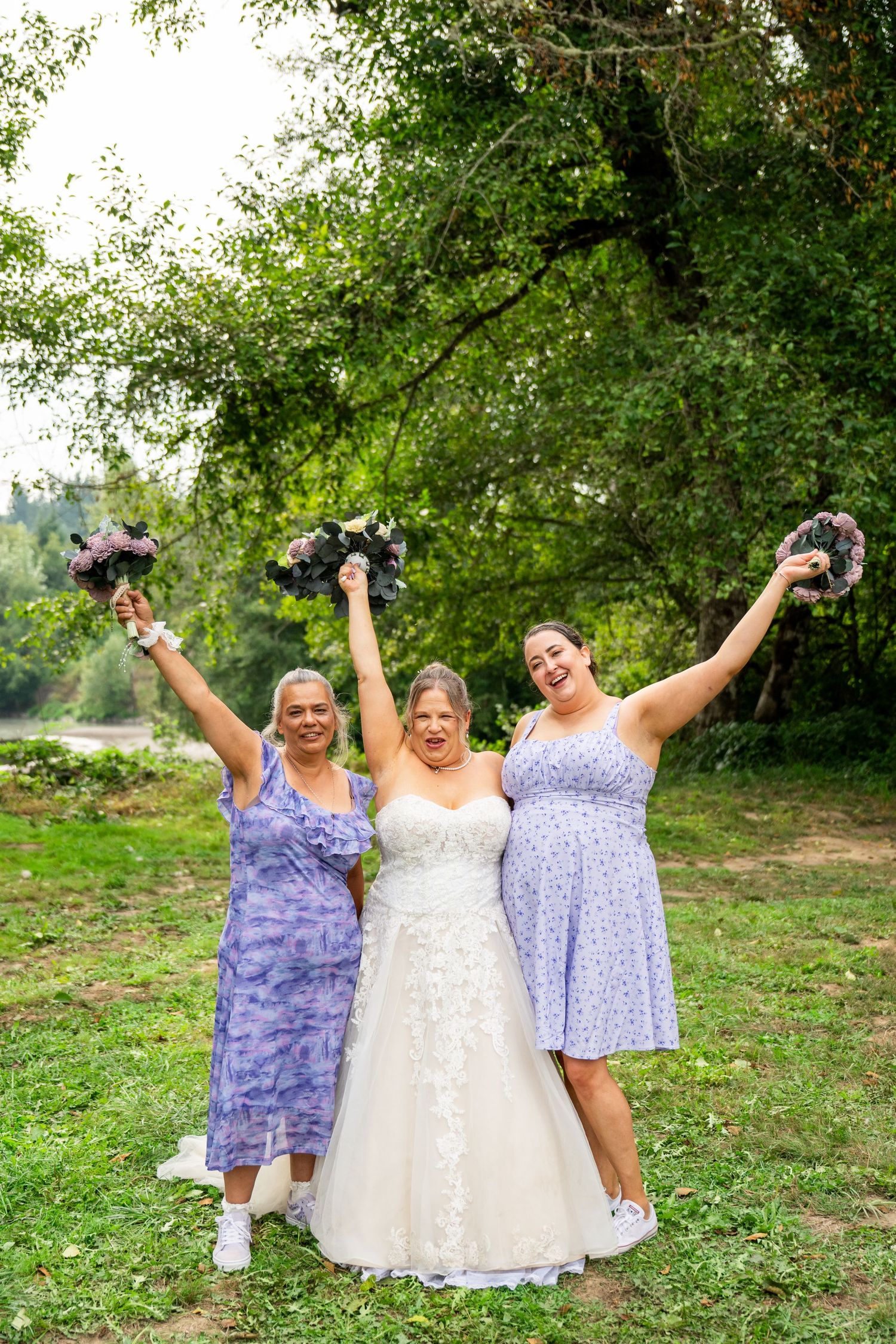 Bride in white lace gown celebrates with two bridesmaids in lavender dresses raising bouquets outdoors.