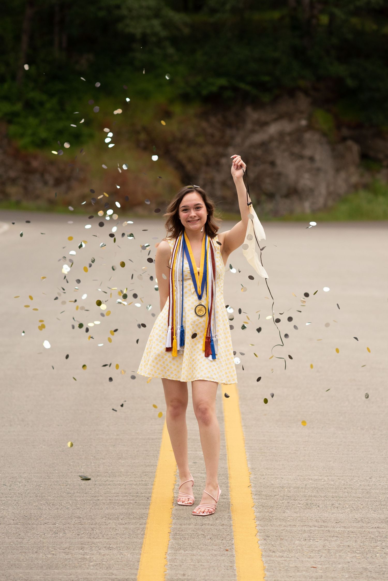 Graduating student in white dress celebrating with confetti on a road wearing honor medals and sashes.