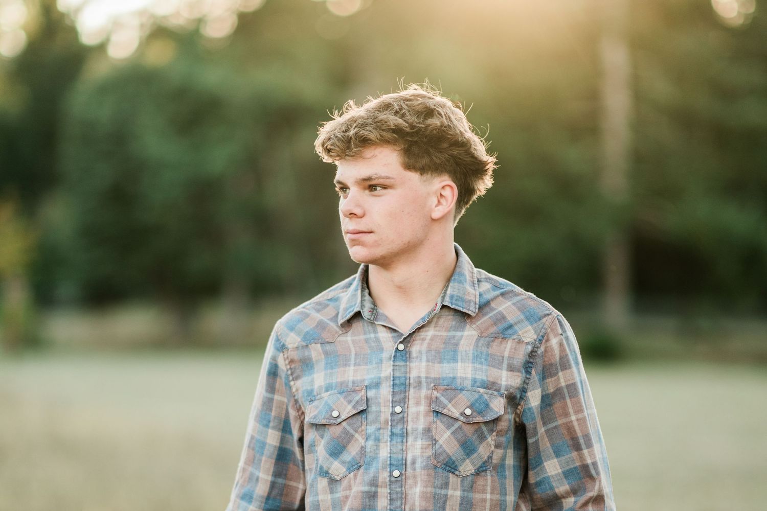 Young man in plaid shirt stands in a sunlit field with trees in the golden hour background.