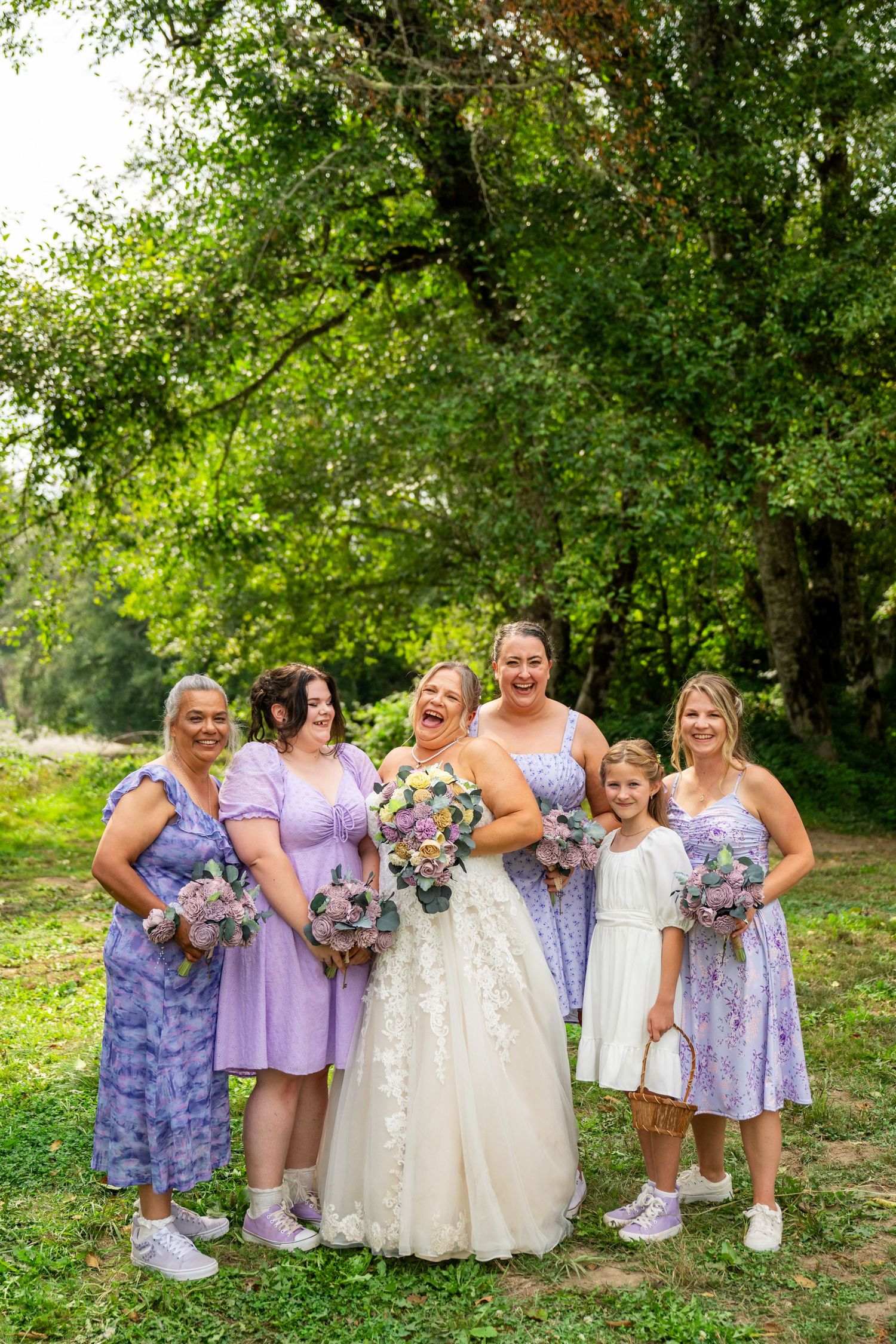 Laughing bride with bridesmaids in lavender dresses and flower girl pose under a large tree.