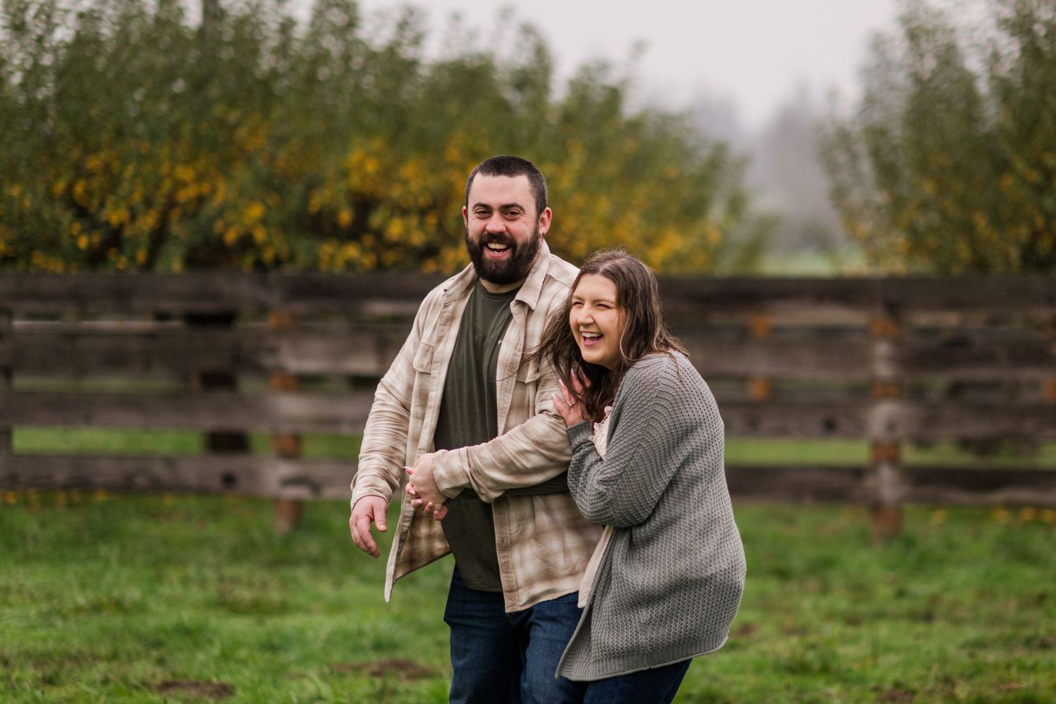Laughing couple playing outdoors near a wooden fence with colorful autumn shrubs in the background on a cloudy day.