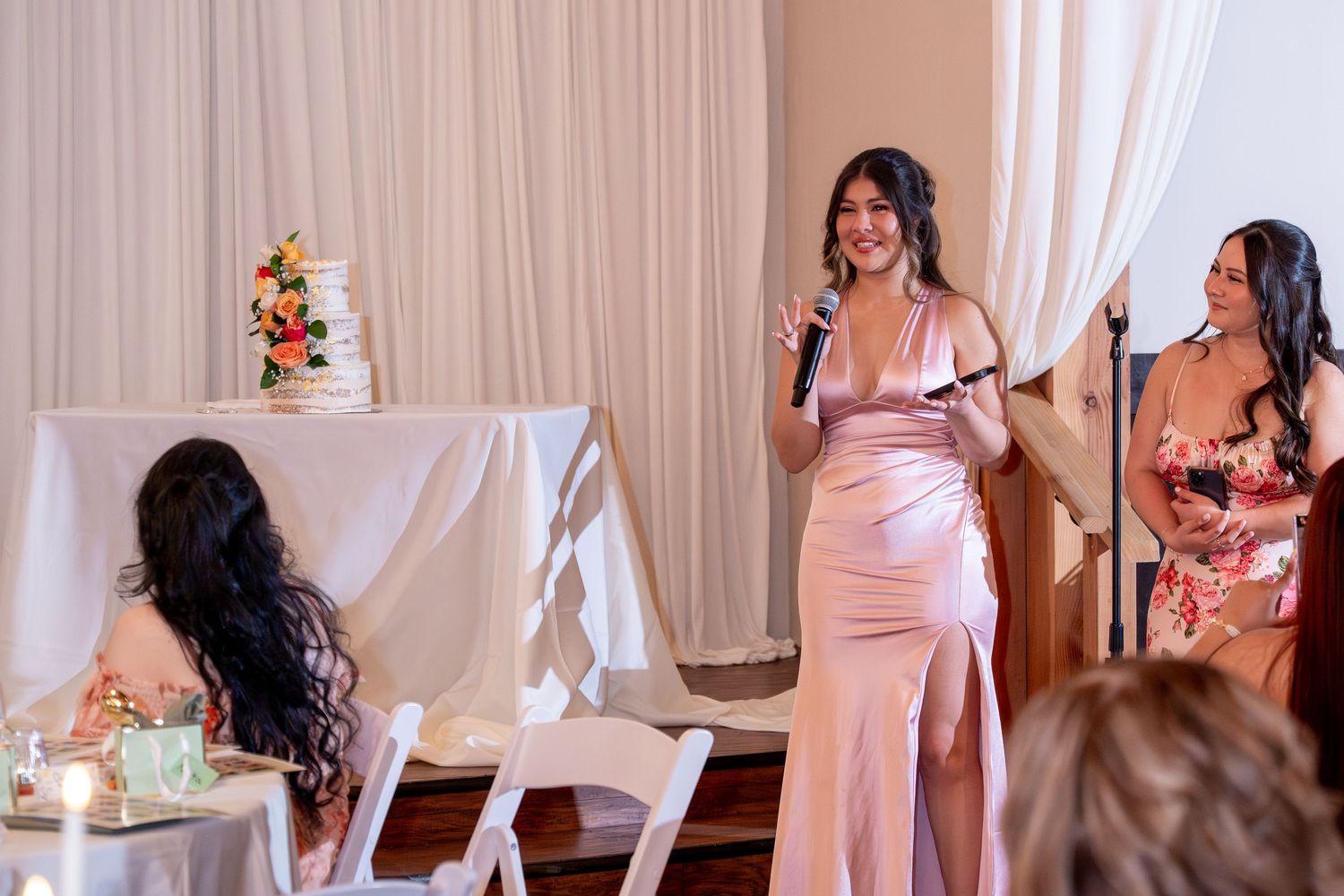 Bridesmaid in pink satin dress gives speech at wedding reception near elegant white cake.