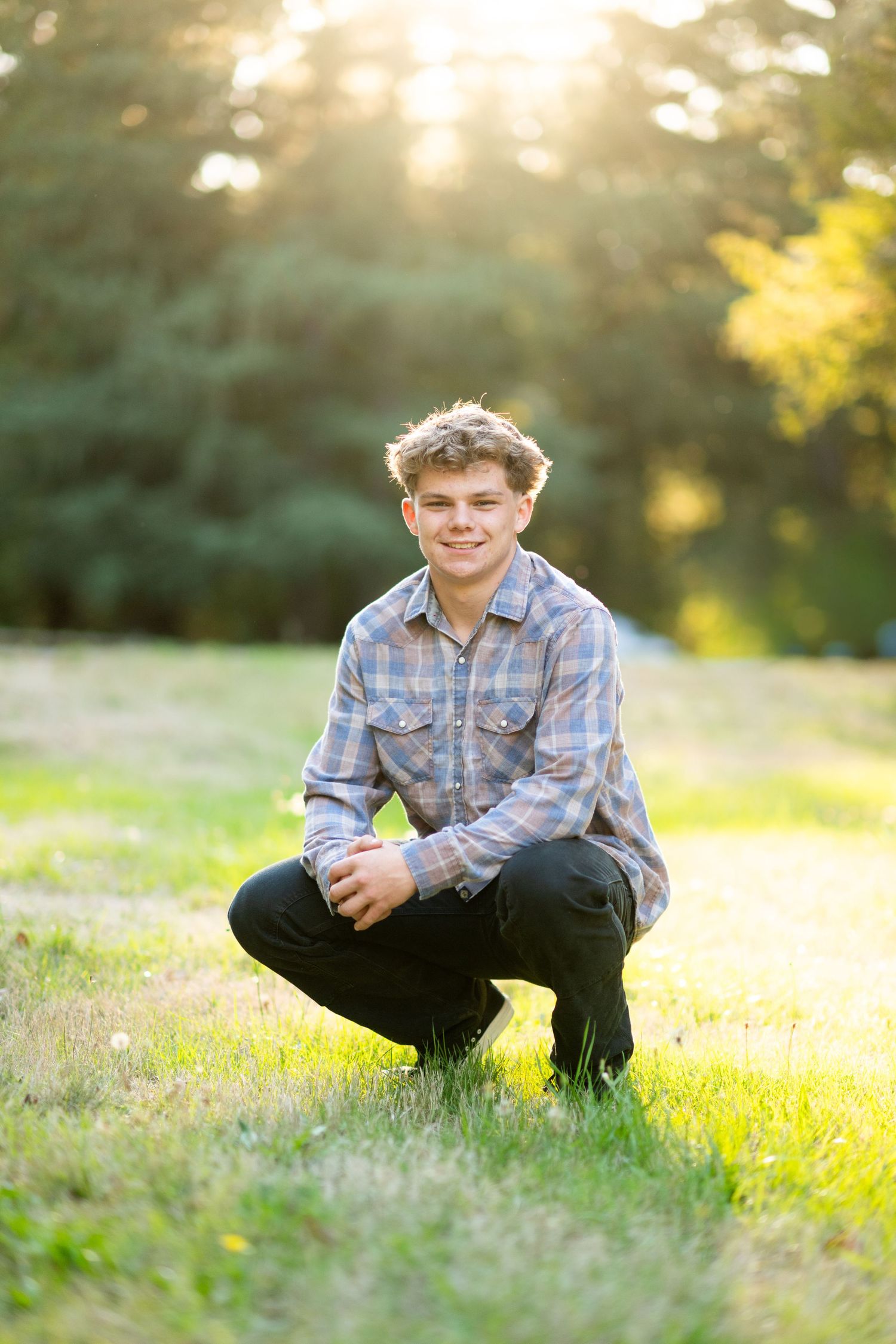 Smiling teenage boy in a plaid shirt crouching on green grass with golden sunlight filtering through trees behind him.