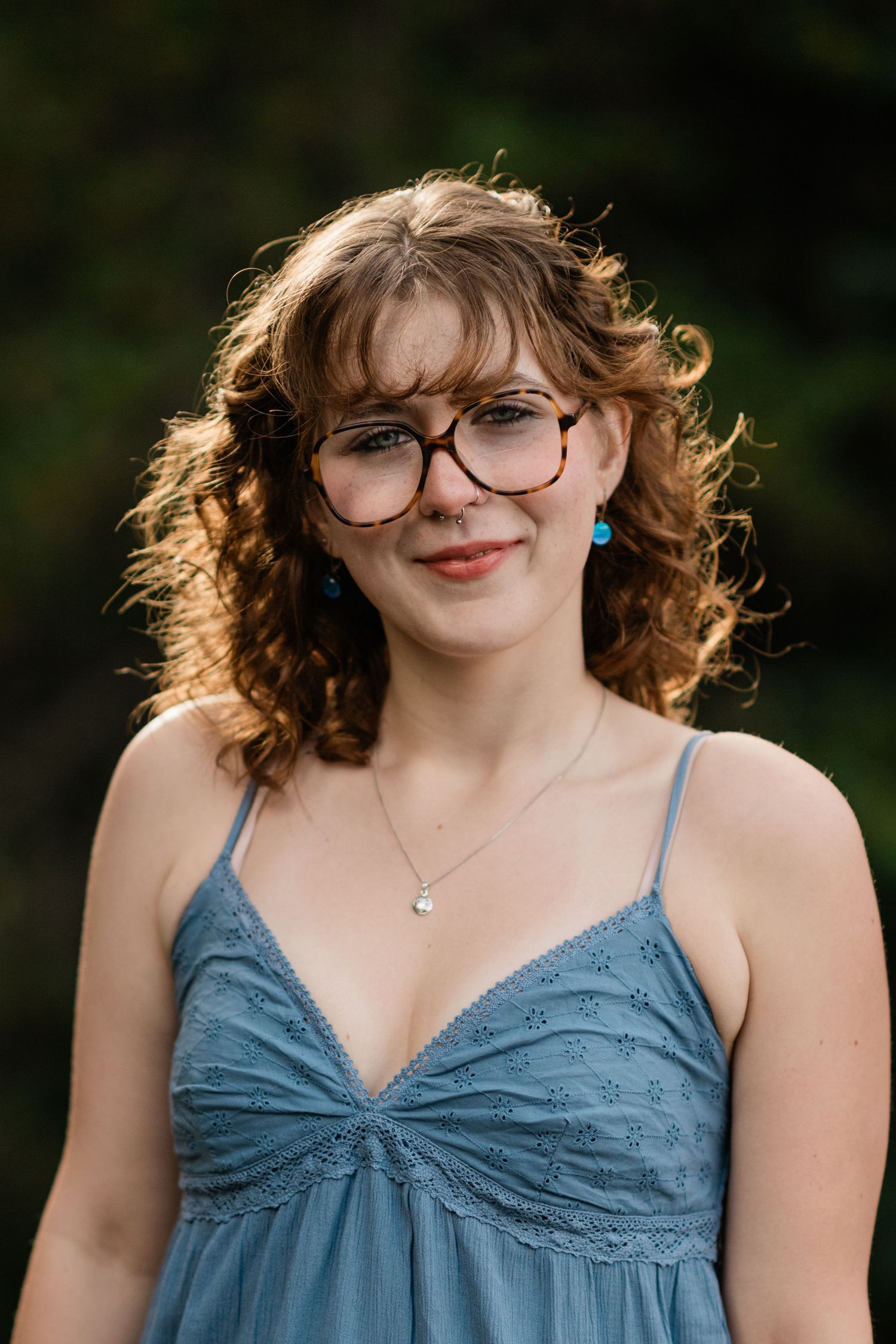 Person in blue dress with curly hair and glasses smiles while wearing turquoise earrings outdoors.