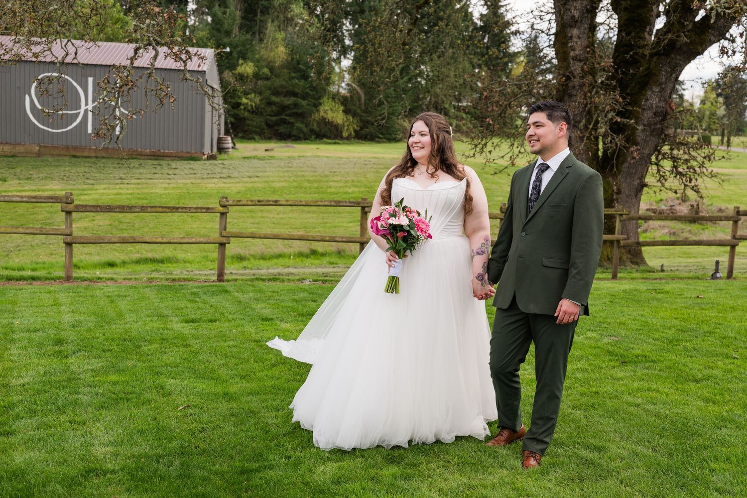 Bride and groom holding hands walking on green grass lawn with rustic barn and wooden fence backdrop.