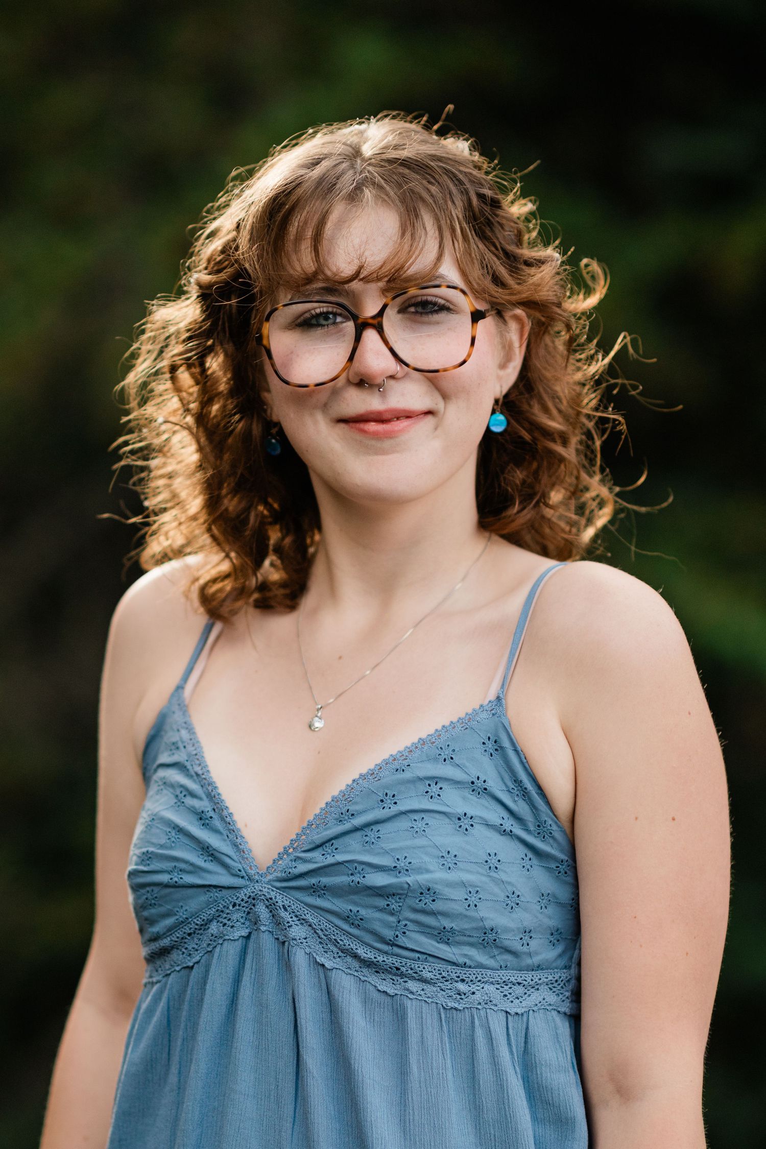 A young woman with curly hair and glasses smiles wearing a blue dress and turquoise earrings outdoors.