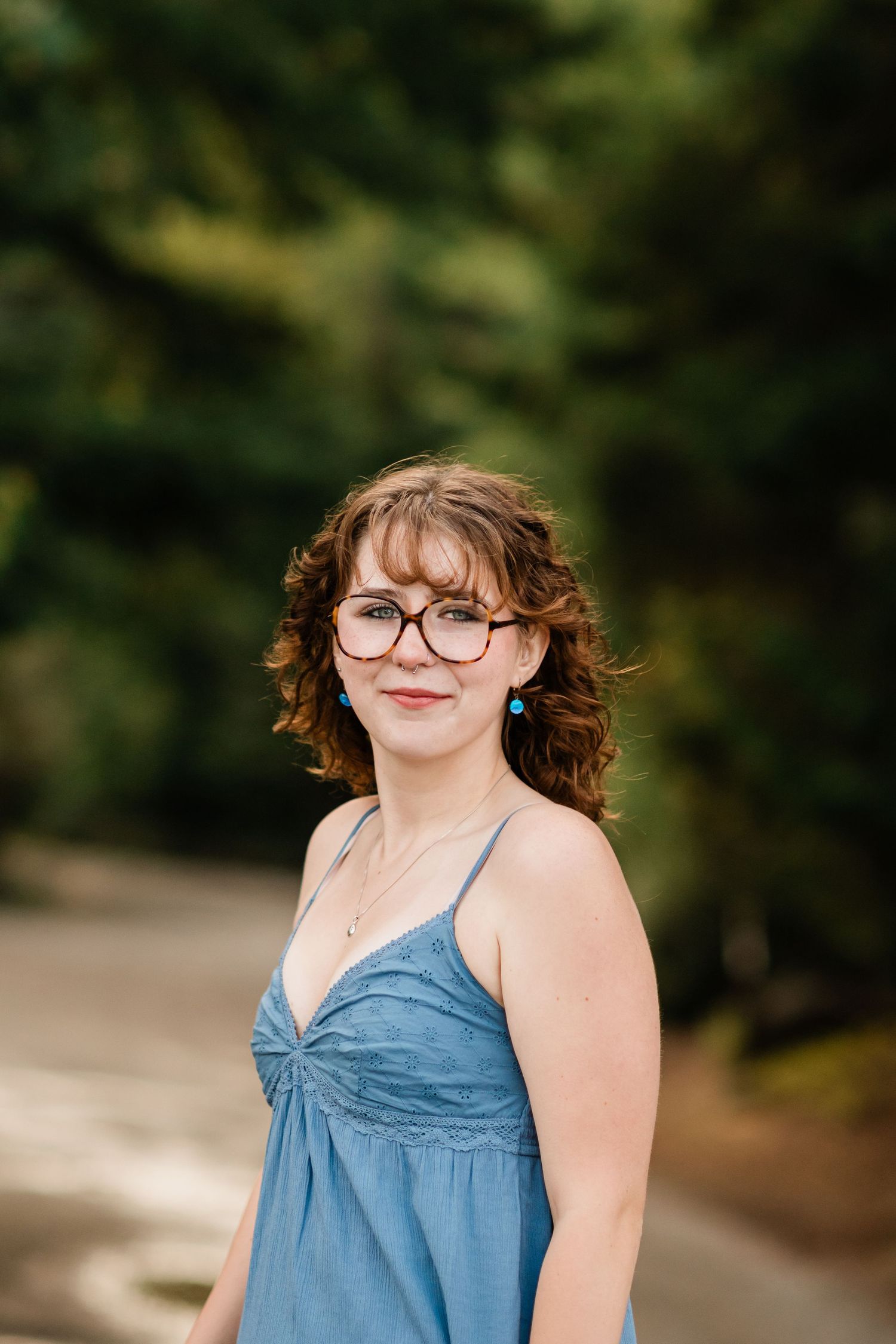 Young woman with curly hair and glasses in a blue sundress standing on a tree-lined road.
