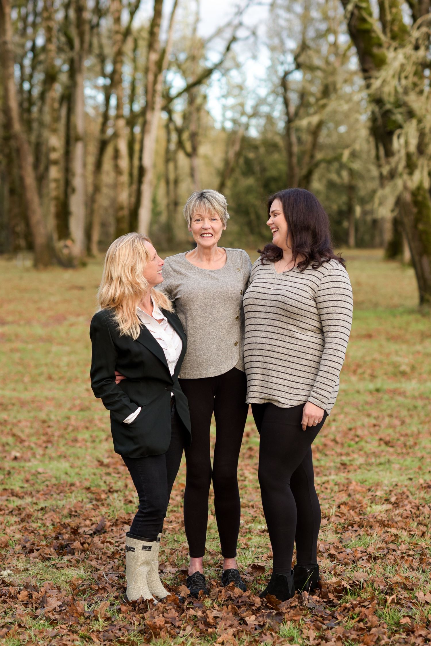 Three women smiling and laughing together while standing among autumn leaves in a peaceful wooded setting.