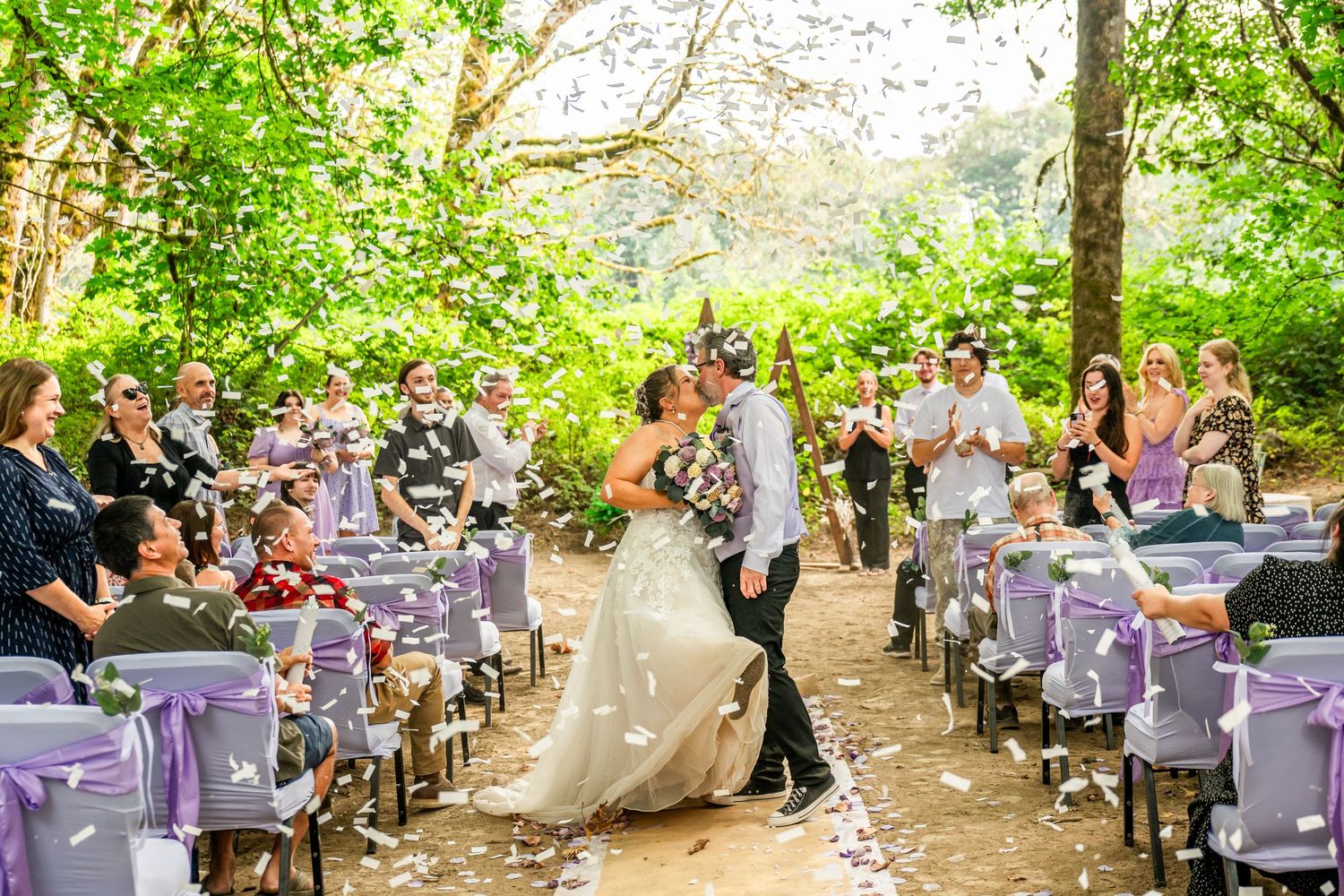 Outdoor wedding ceremony setup with white chairs and purple decorations in a wooded setting.