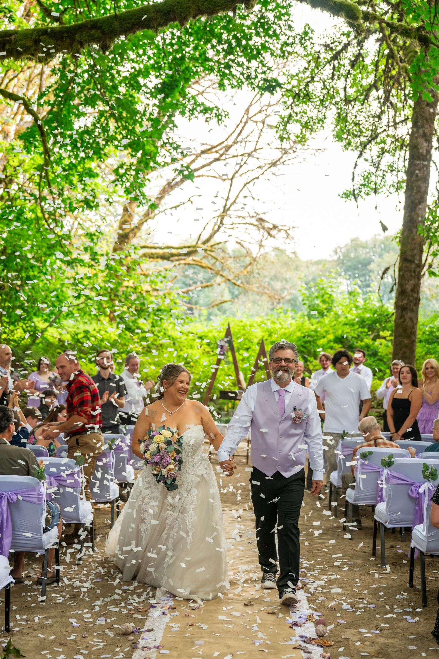 Newlyweds walk back down aisle as guests toss white confetti at outdoor woodland wedding ceremony.