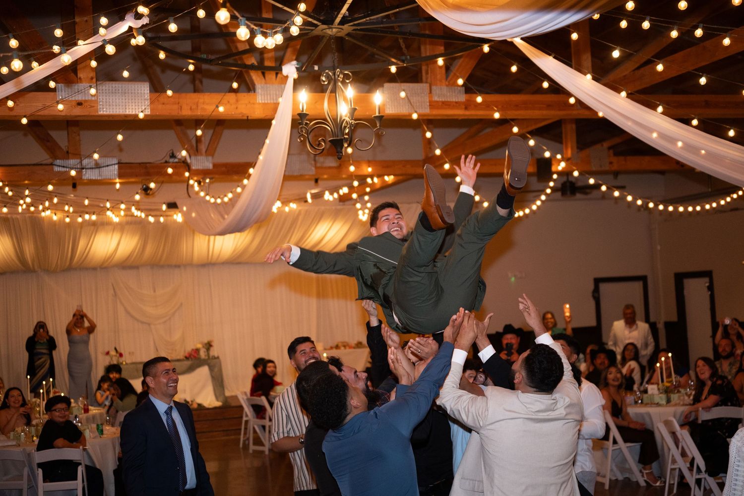 Groom tossed in the air by groomsmen during lively wedding reception in decorated banquet hall with string lights.