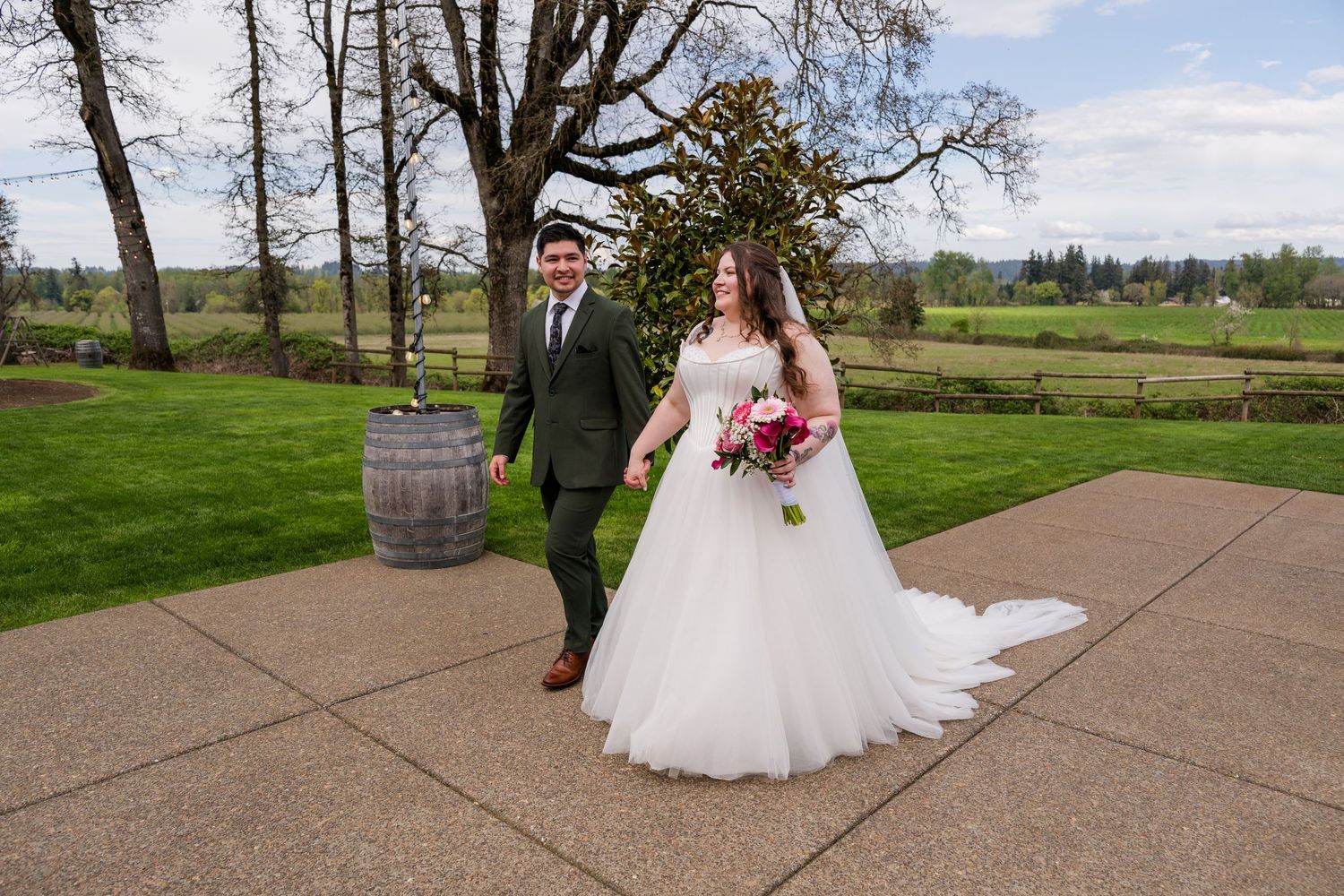 Bride in white ballgown and groom in dark suit walk hand in hand on a paved path at an outdoor wedding venue.