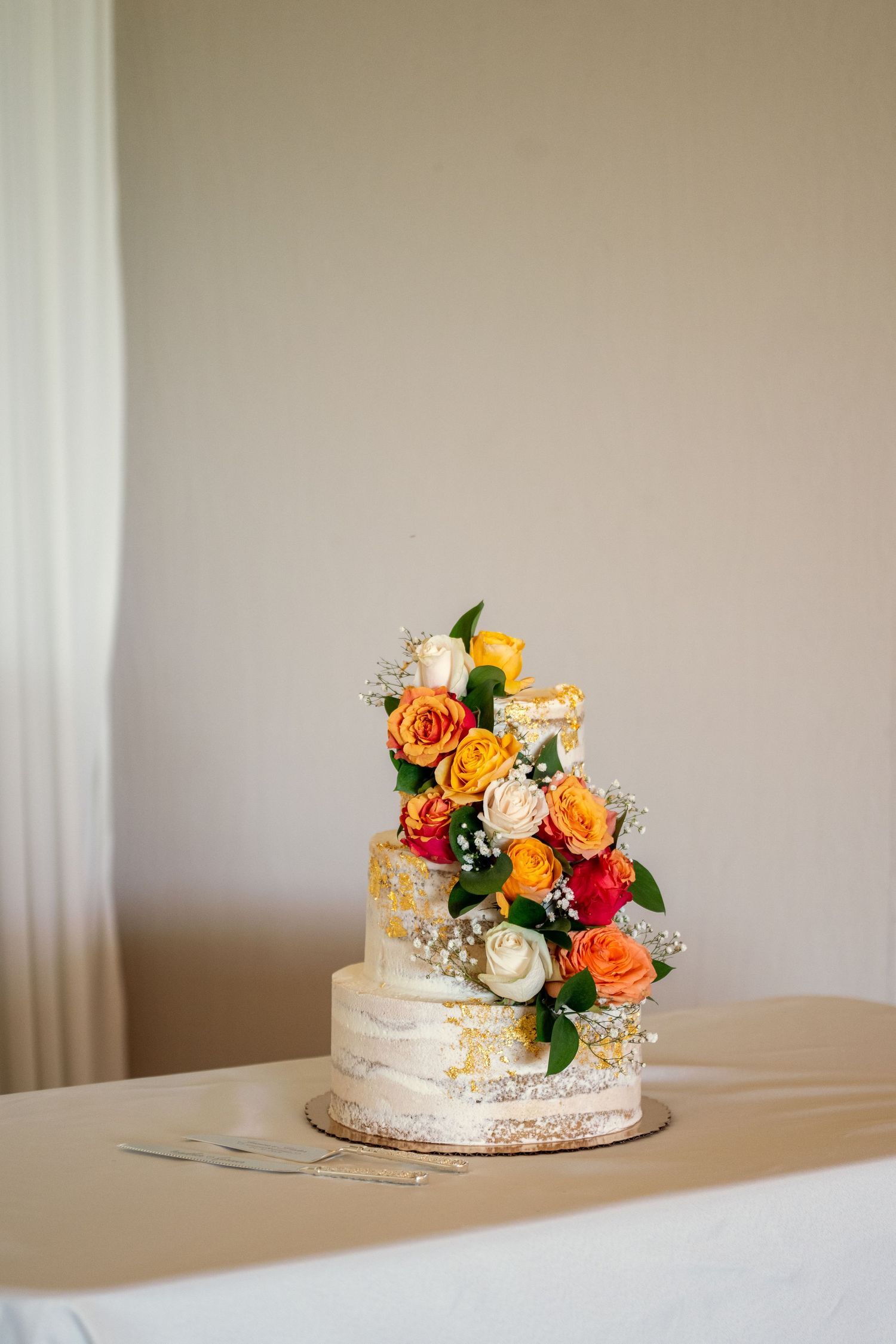 Three-tier semi-naked wedding cake adorned with vibrant orange, red, and white roses with gold leaf accents on a white table.