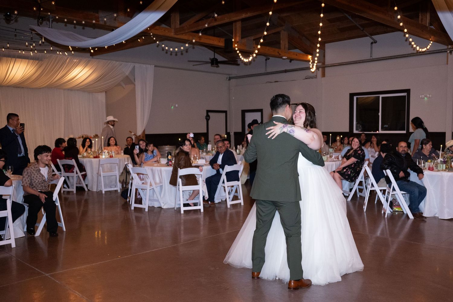 Bride and groom share their first dance at a romantic wedding reception with guests seated at white-draped tables.