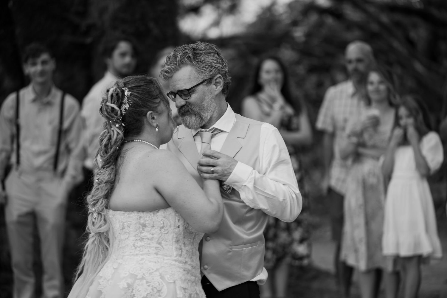Black and white photo of bride and groom sharing first dance at outdoor wedding reception with guests watching.