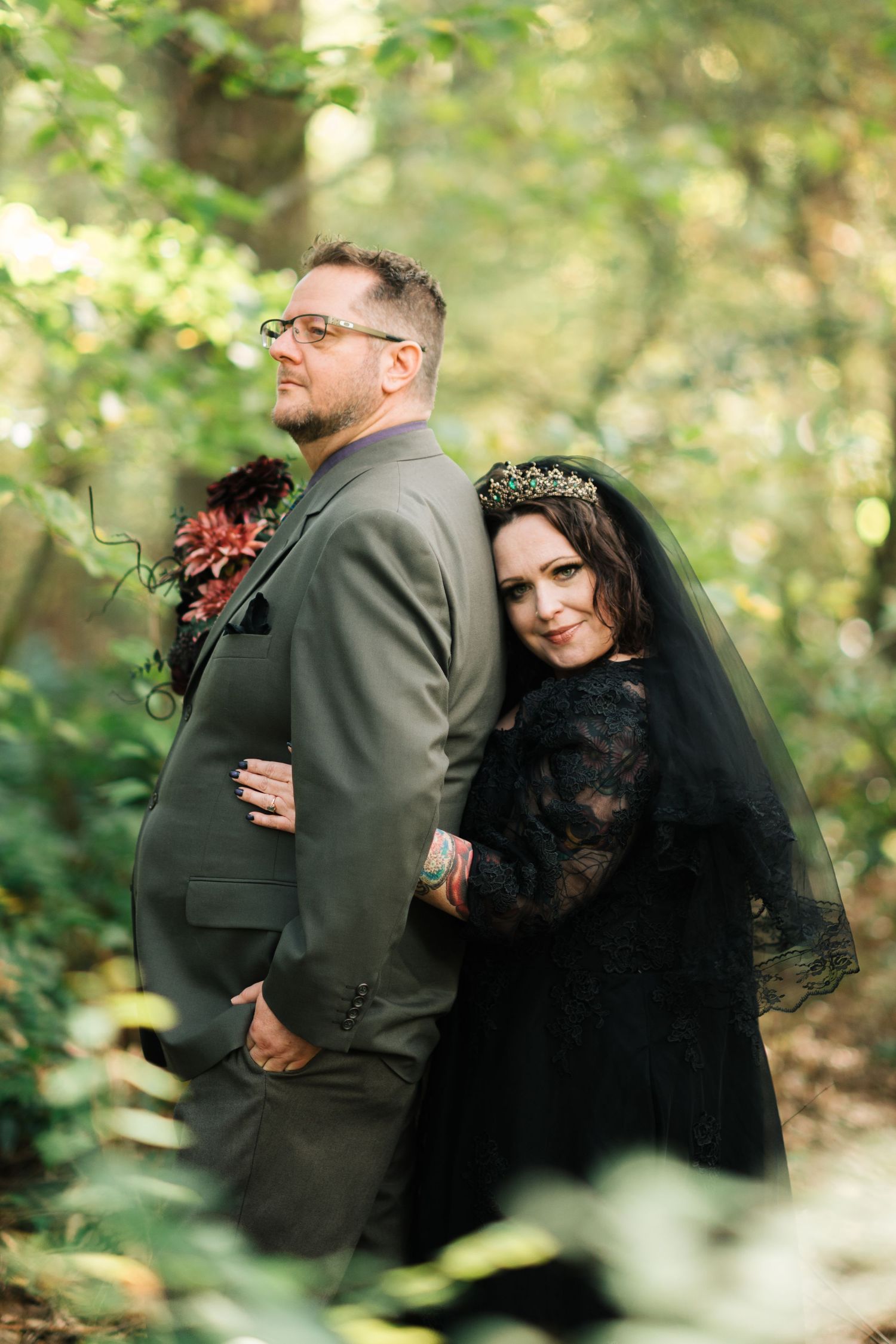 Gothic wedding couple in forest, bride in black lace gown with crown veil leans against groom in olive suit.