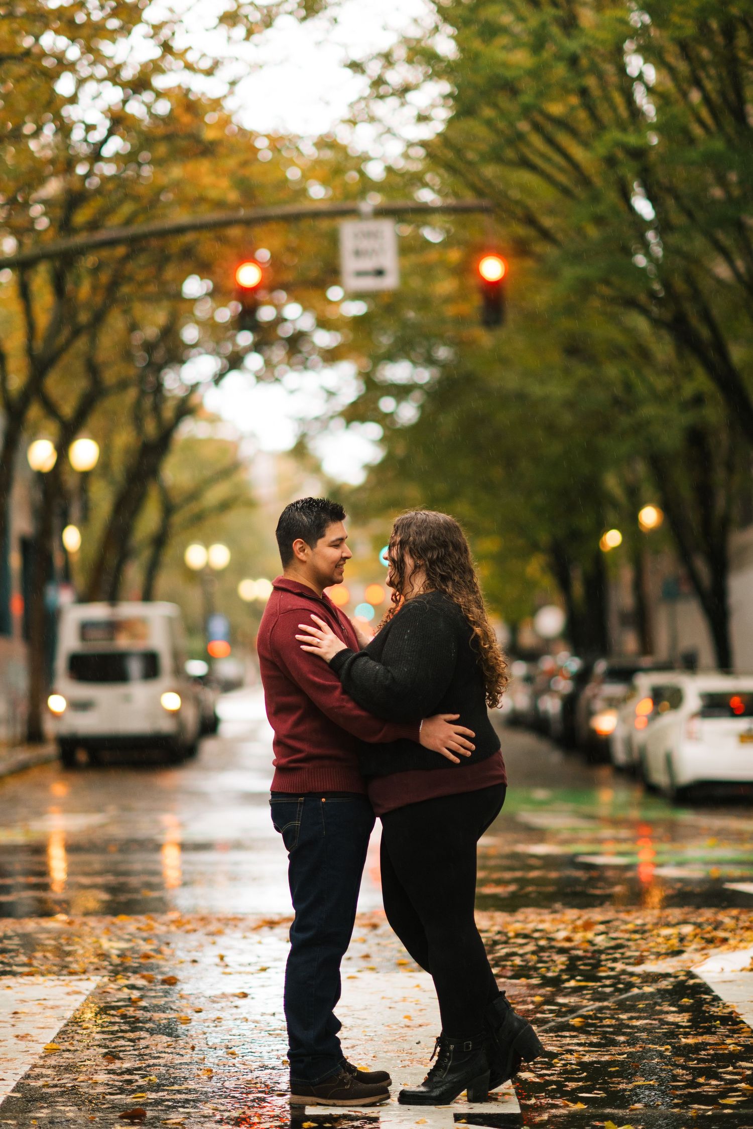 Couple embracing romantically on a rain-soaked street lined with fall foliage during an evening engagement shoot.