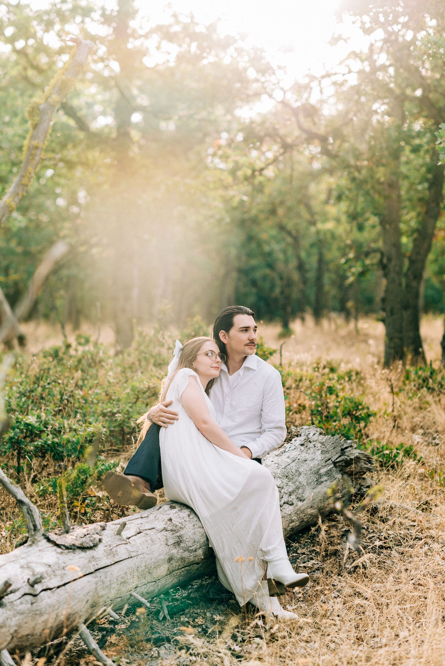 A couple in white clothing sitting together on a fallen log in a sun-dappled forest.