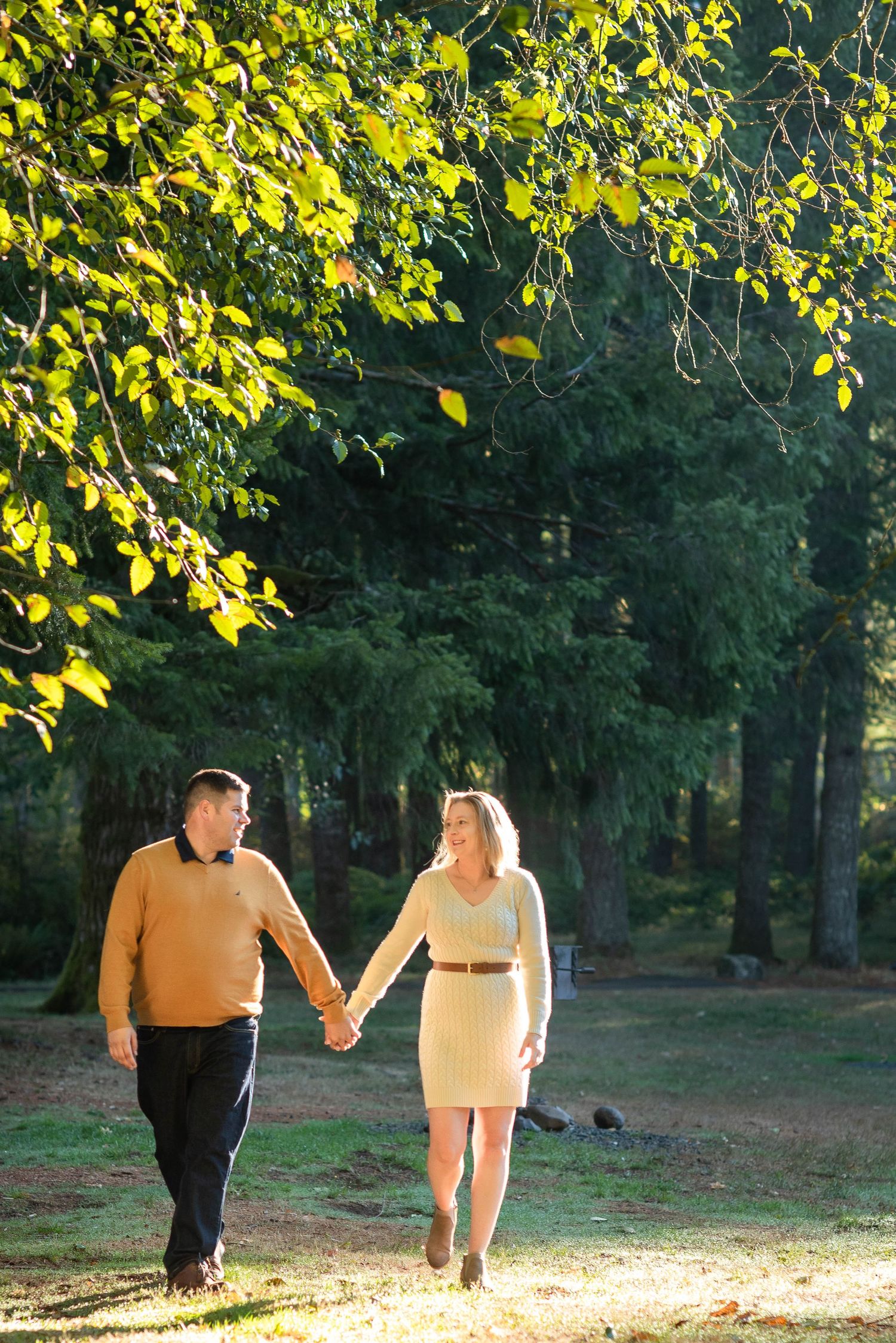 Couple holding hands while walking through sunlit forest path in casual attire.