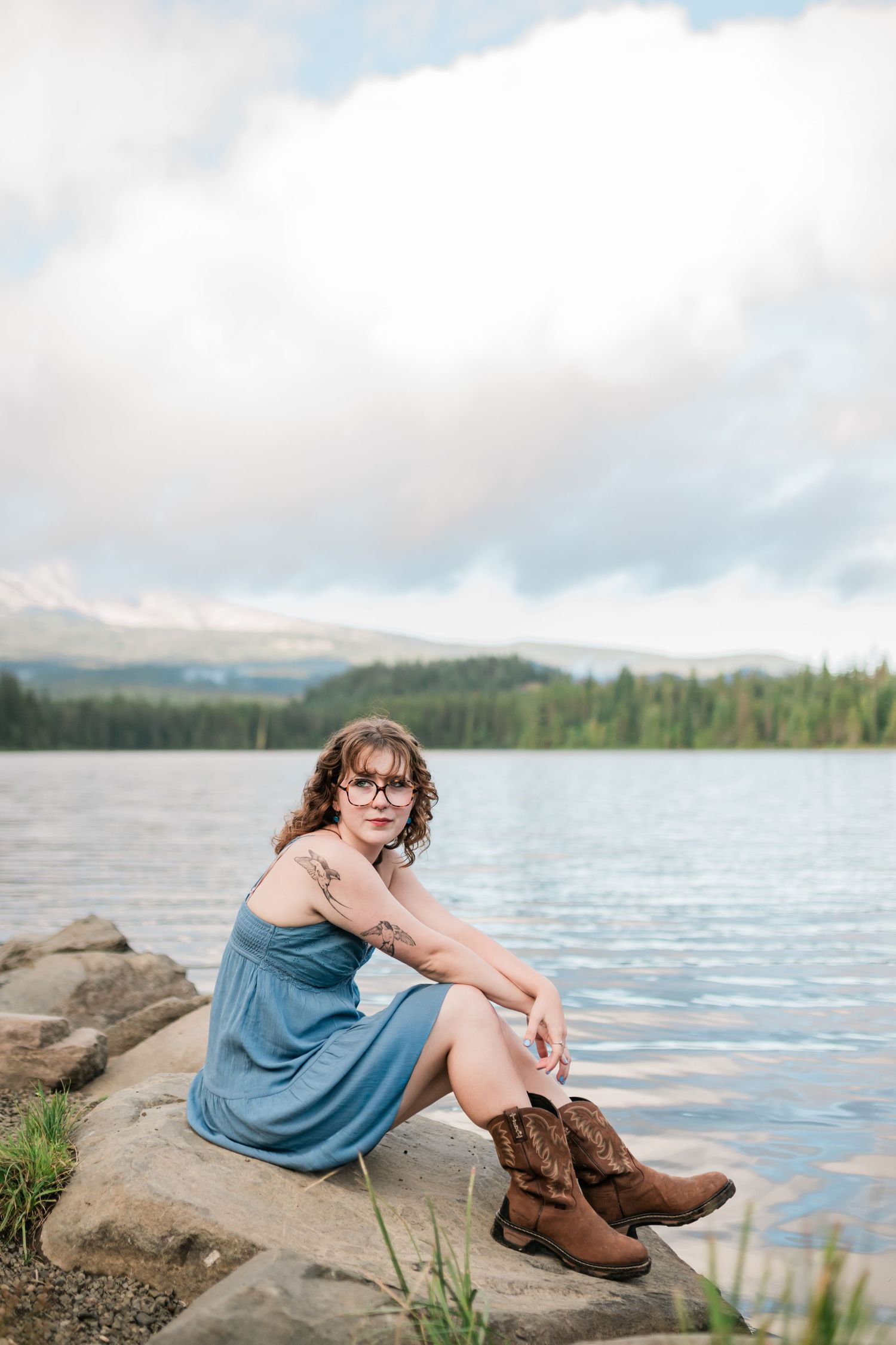 Woman in blue dress and cowboy boots sits on rocks by a serene mountain lake under cloudy skies.