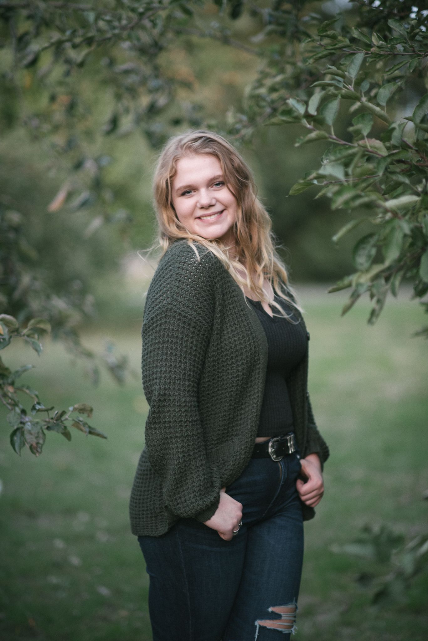 Smiling blonde teen in dark green knit cardigan posing among green apple tree branches outdoors.