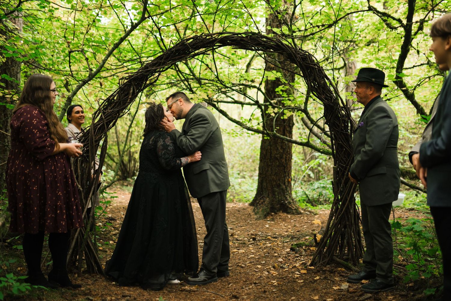 People gather in a lush green forest setting for an outdoor ceremony or event under tree canopy.