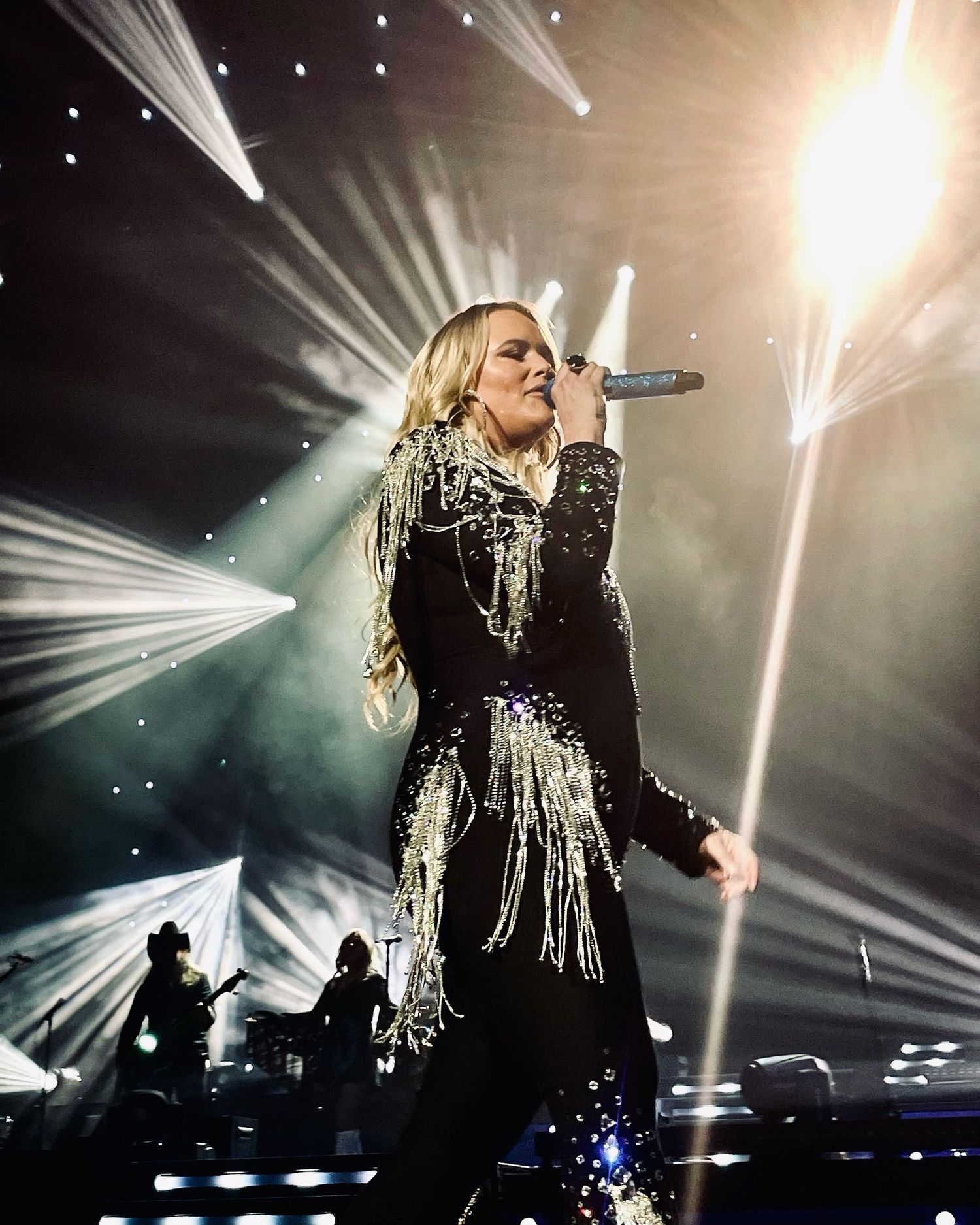 Female performer in black fringed outfit sings on stage with dramatic concert lighting behind her.
