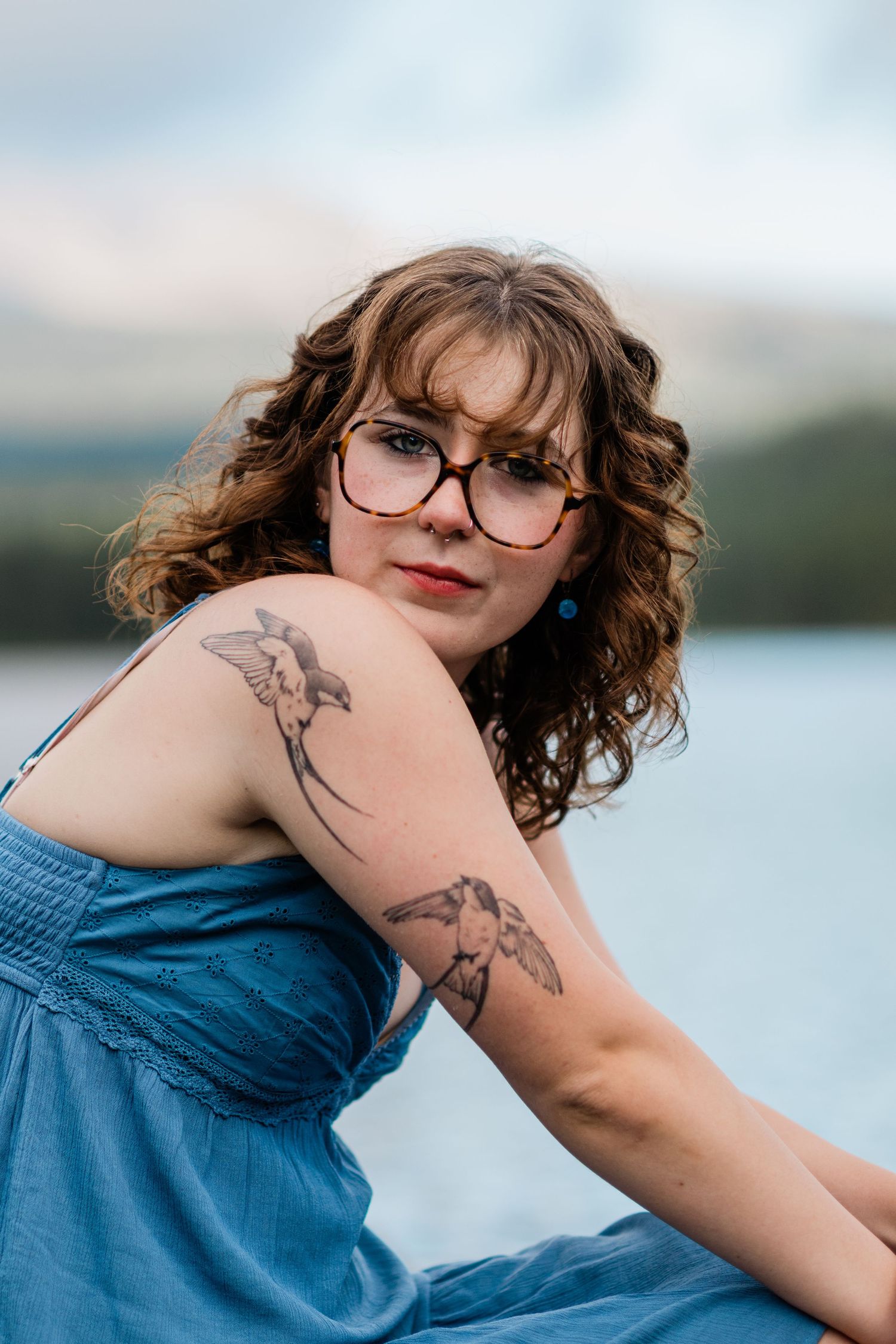 Person in blue dress with bird tattoos poses near water wearing glasses and showing curly hair.