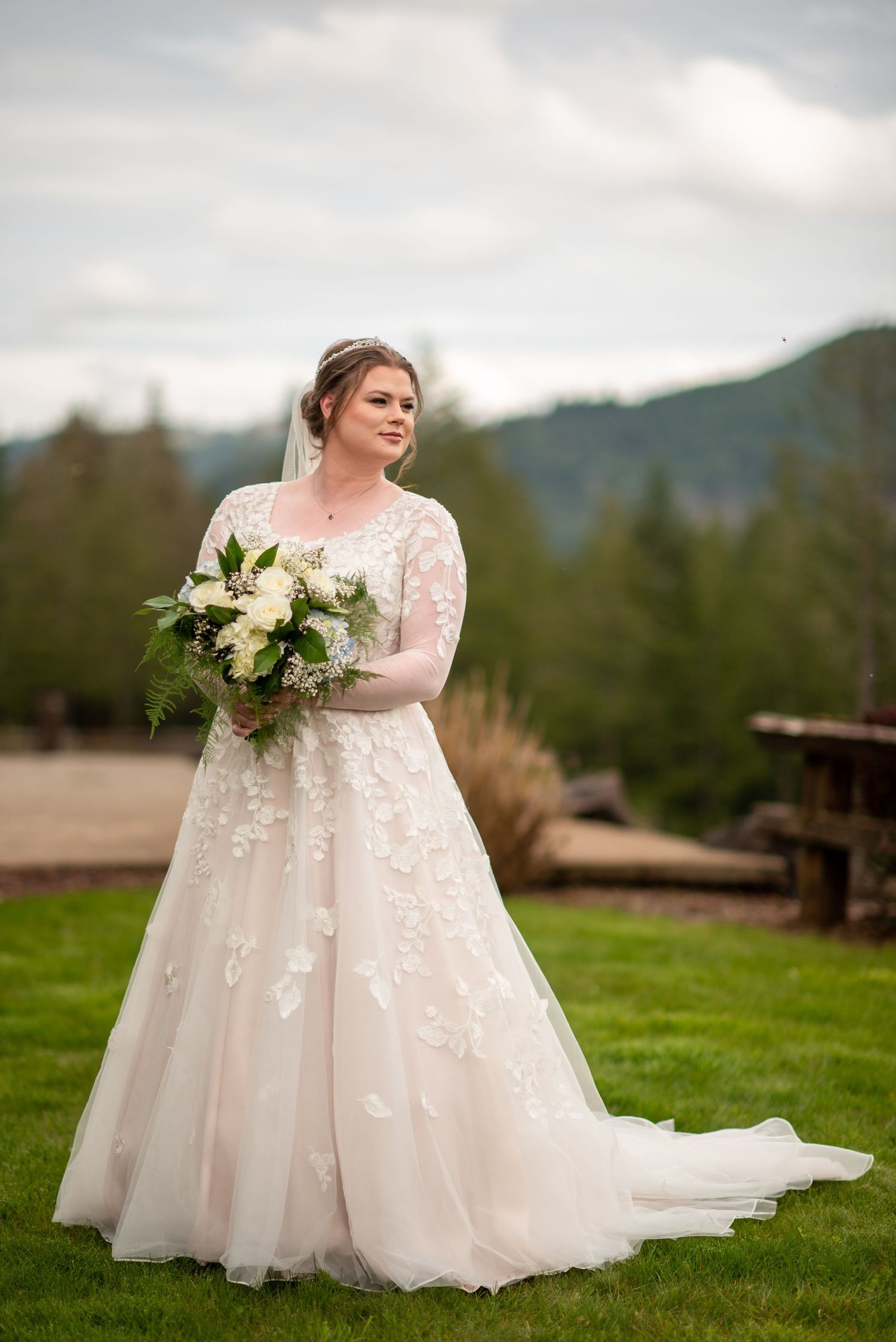 A bride in a lace-detailed wedding gown holds a white bouquet against a mountain backdrop.