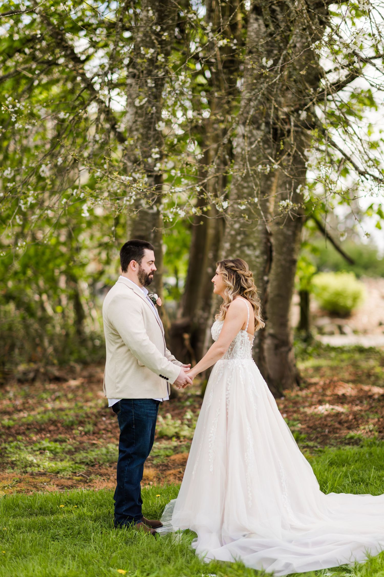 Bride and groom hold hands and smile at each other beneath a blooming tree in a green garden setting.