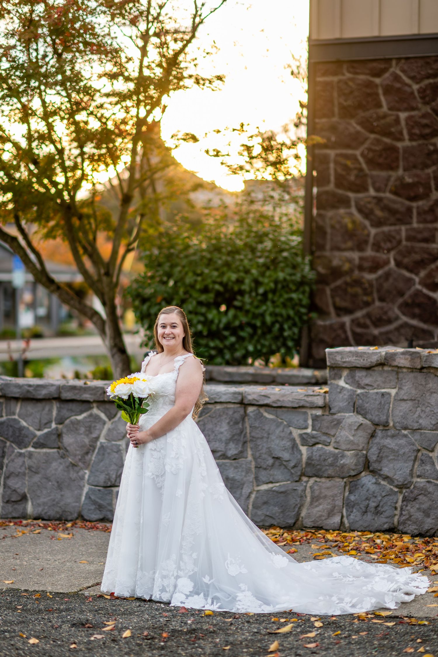 Bride in white lace gown holds yellow and white bouquet at golden hour beside a stone wall outdoors.