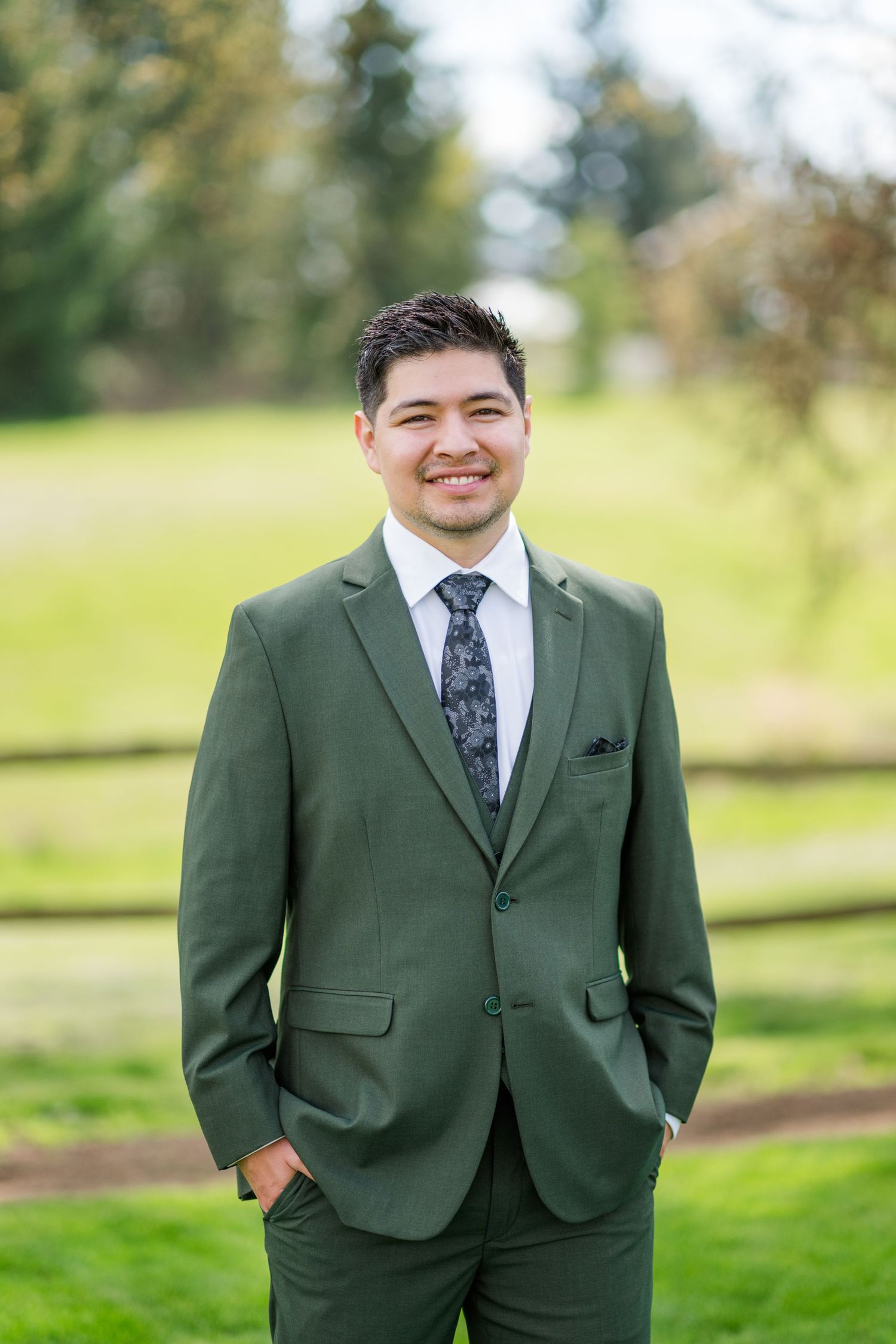 Smiling man in olive green suit with patterned tie standing outdoors in a park setting.